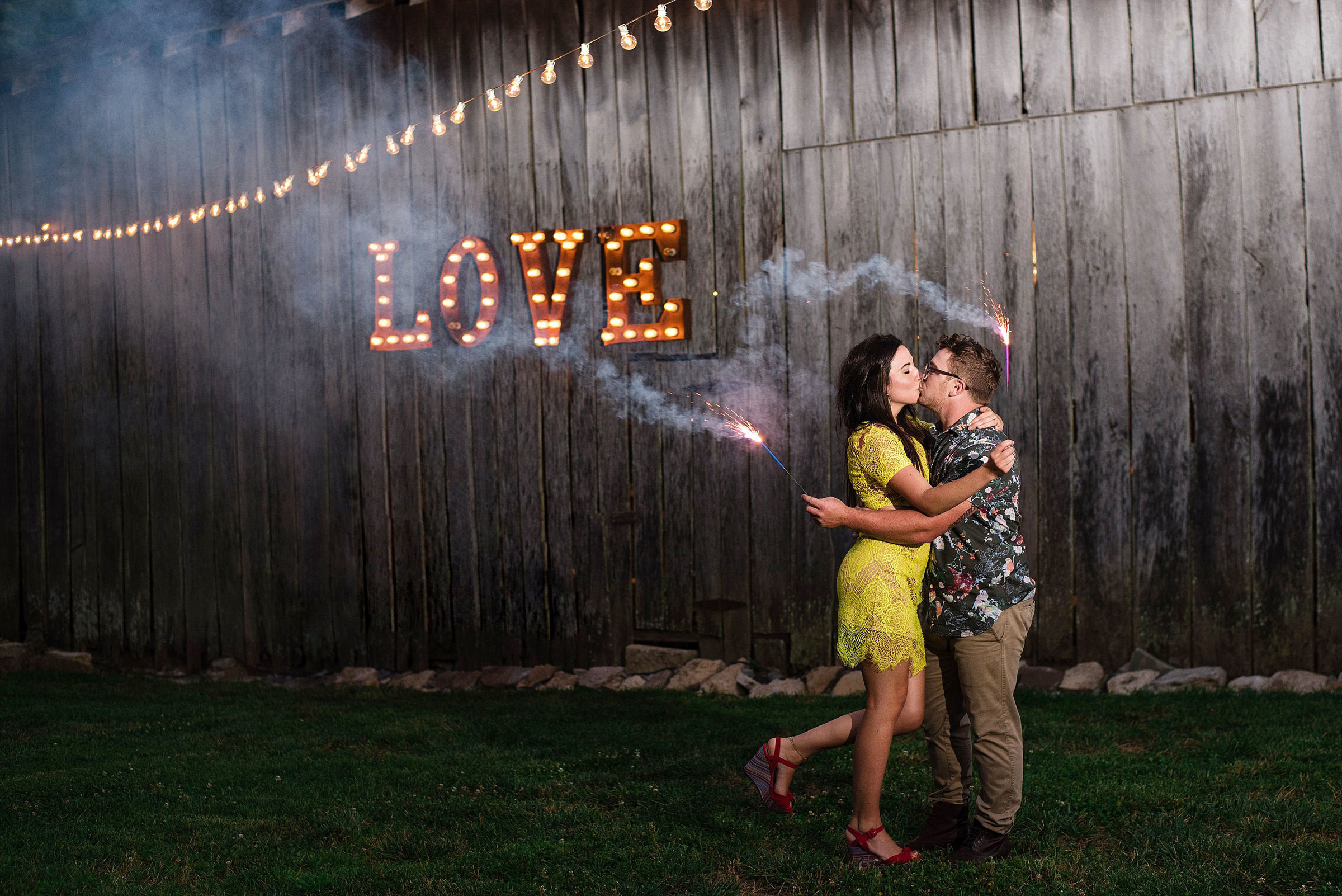Couple holding sparklers and kissing in front of the Love light up letter sign on the barn at Drakewood Farms