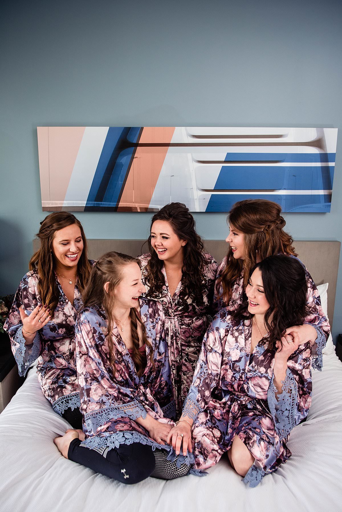 Bride with her girlfriends  in purple and pink robes sitting together on a bed