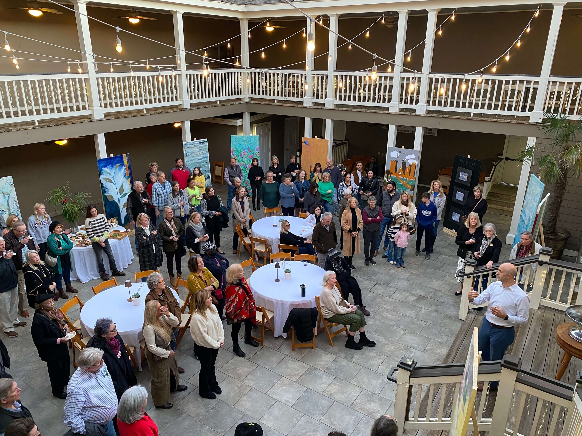 A large group of people gathers in a spacious outdoor courtyard with string lights, round tables, and colorful artwork on display. A speaker addresses the crowd from the bottom of a staircase.