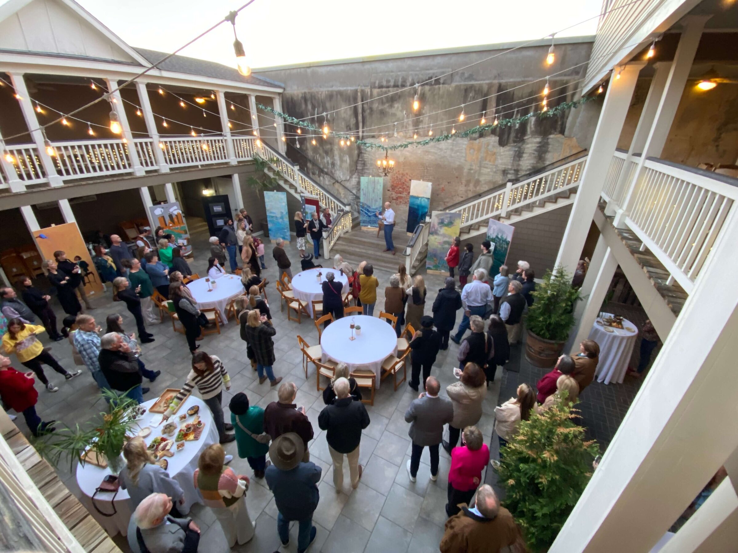 A man stands on stairs speaking to a gathered crowd at an art exhibit on the stairs in an open couryard. Large paintings are displayed around him, and people listen and observe from below.