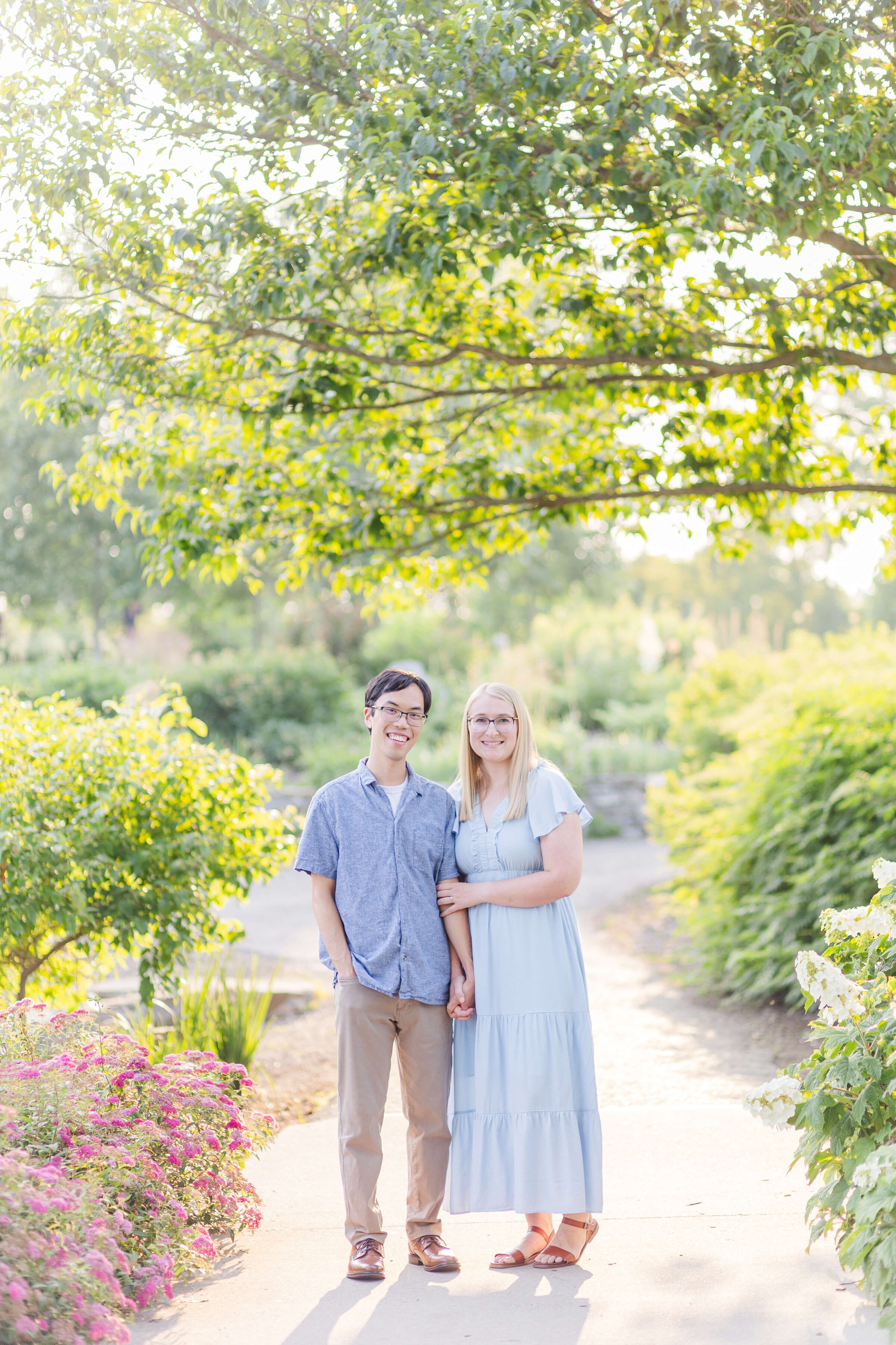 A man holds hands with his fiancee who is wearing a blue dress. It's a beautiful summer evening as the sun glows through trees.