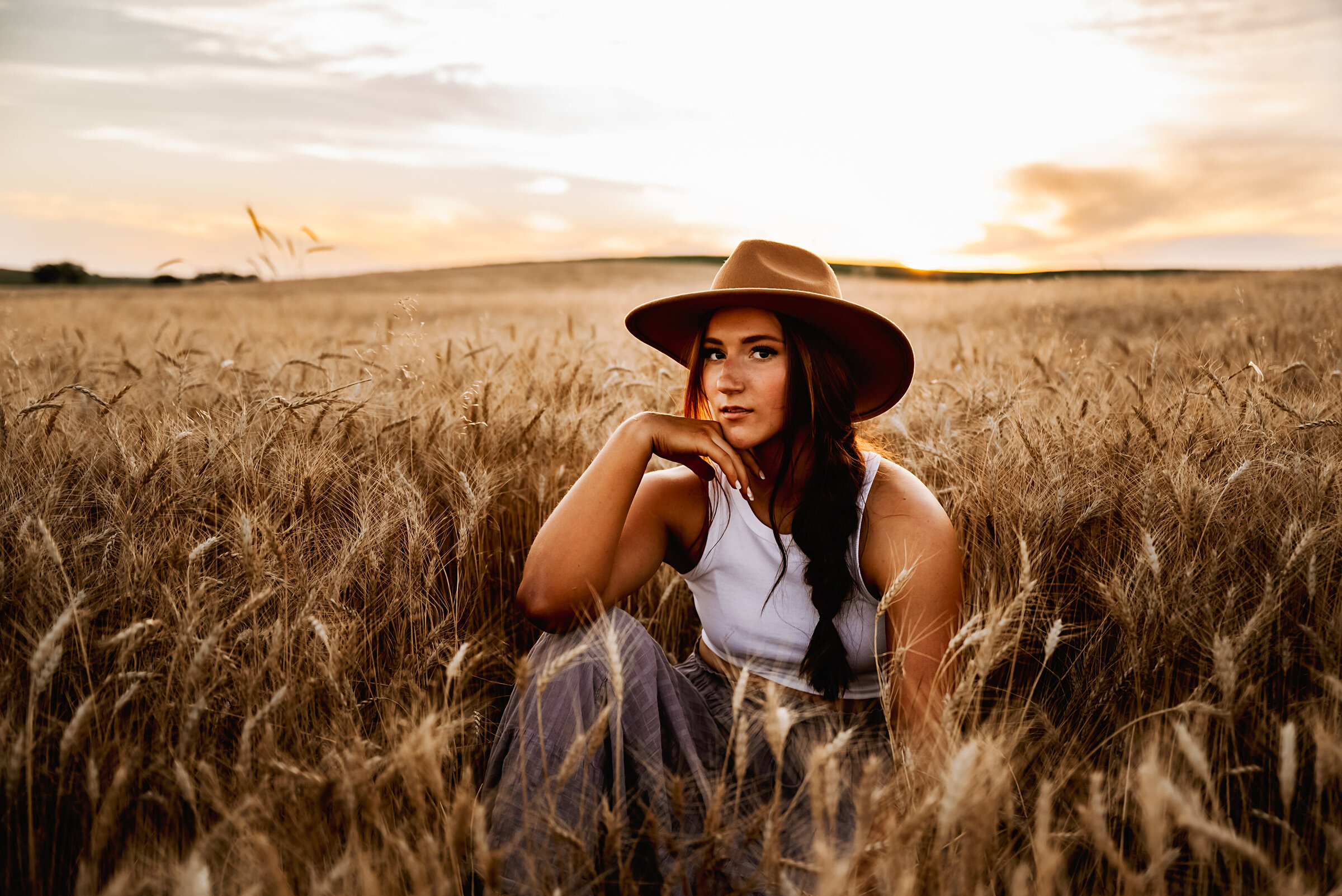 senior girl sits in wheat field with colorful sunset and wide brim hat