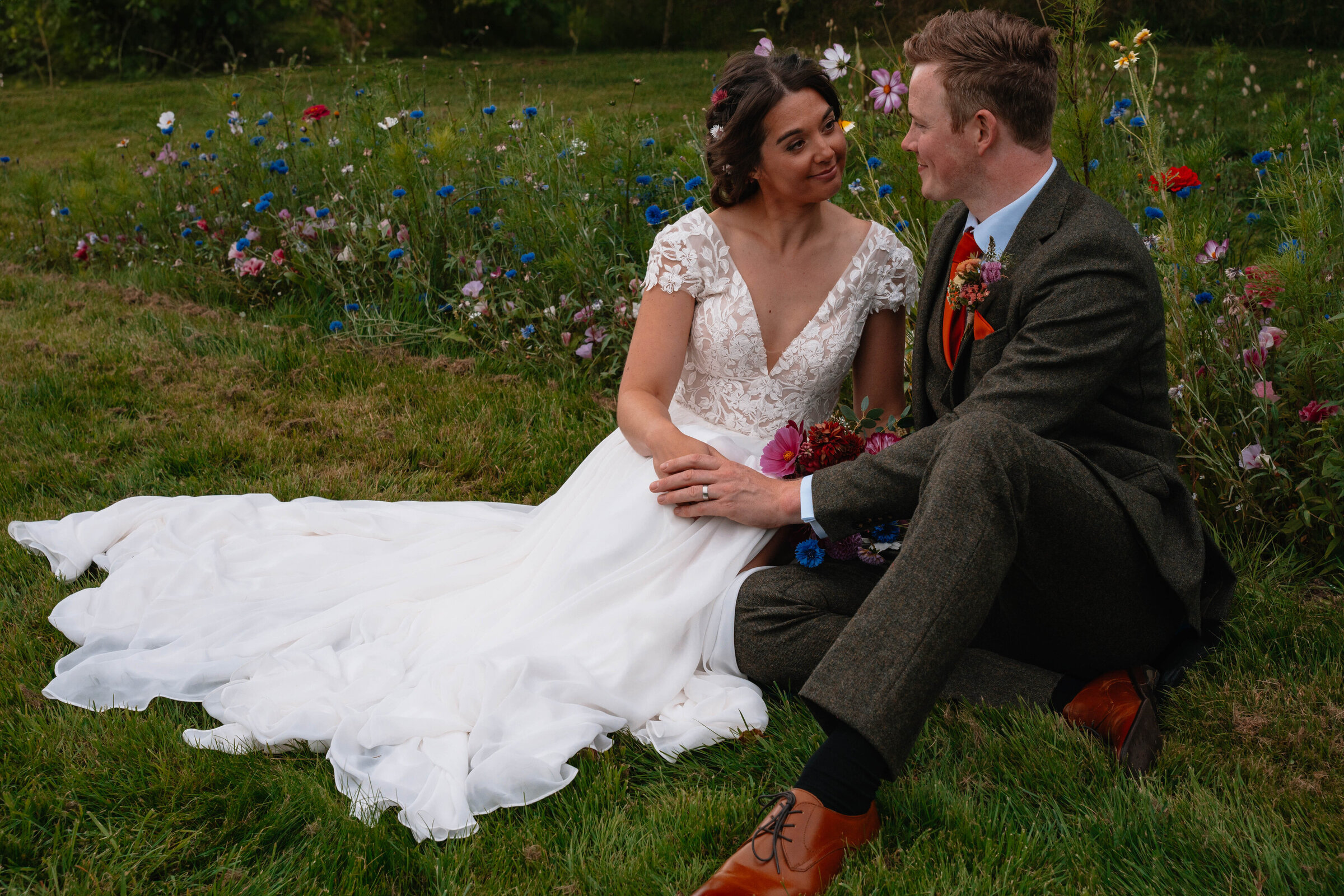 Bride and Groom in wedding attire seated close together on the grass with wildflowers in the background