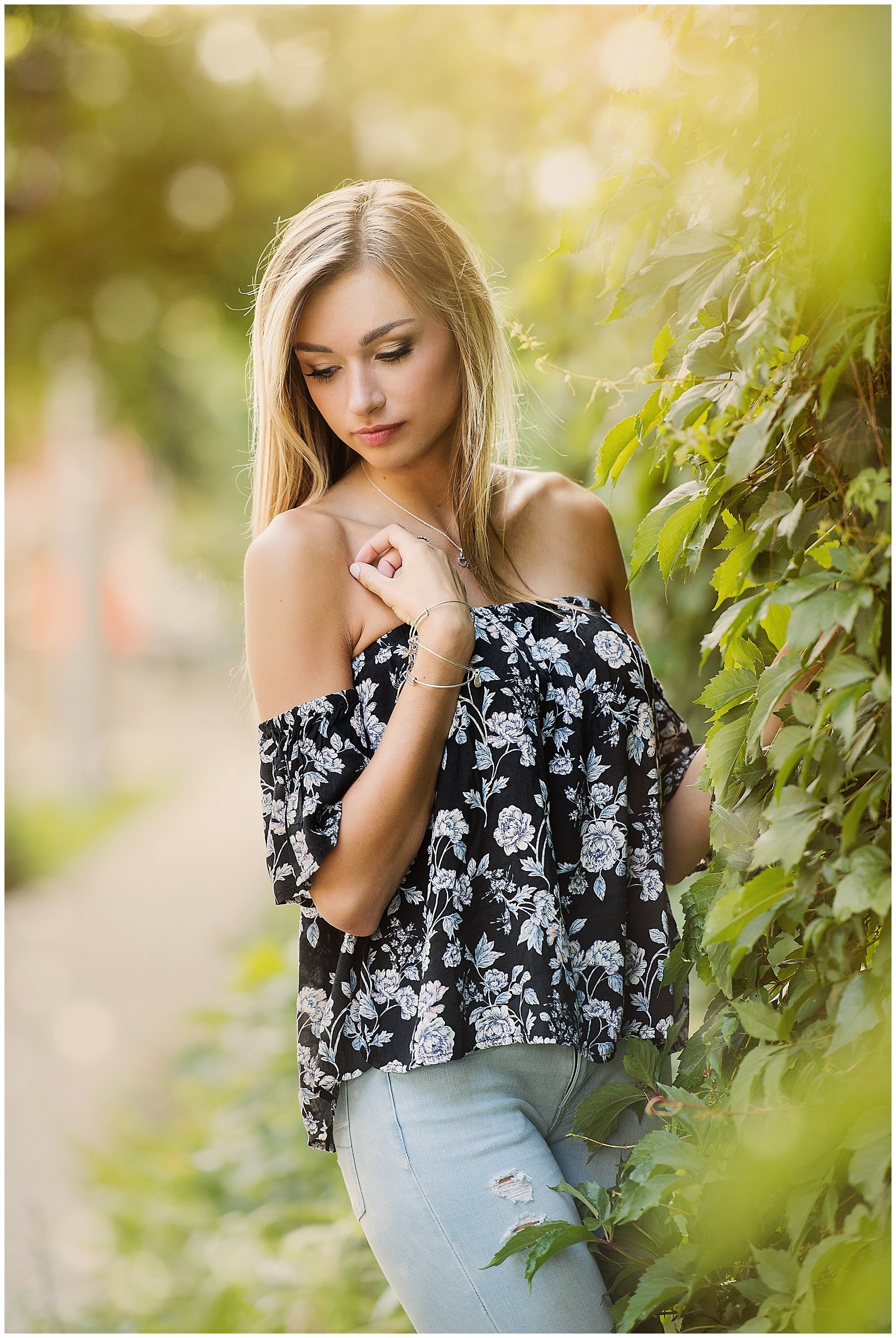high school senior photo of girl with flower shirt and jeans against ivy wall with sun flare