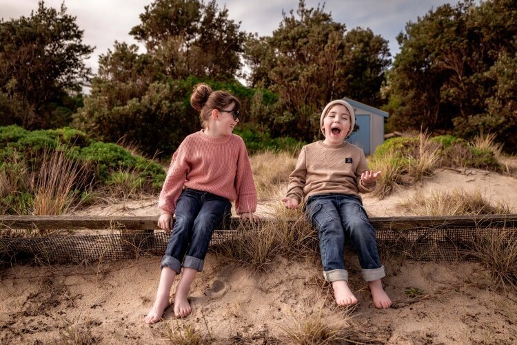Two children in jeans sitting at beach laughing by Melbourne Family Photographer Kate Cashin