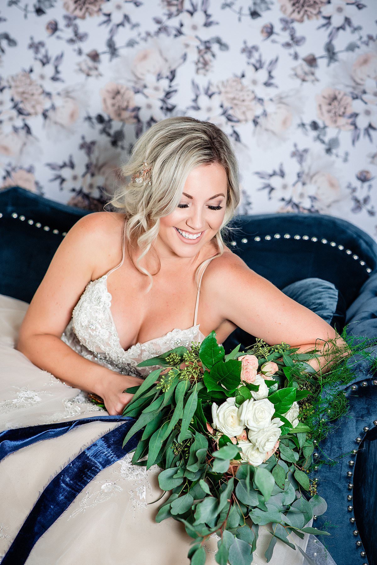 Bride sitting on a blue velvet couch holding her greenery heavy bouquet with floral wallpaper behind her