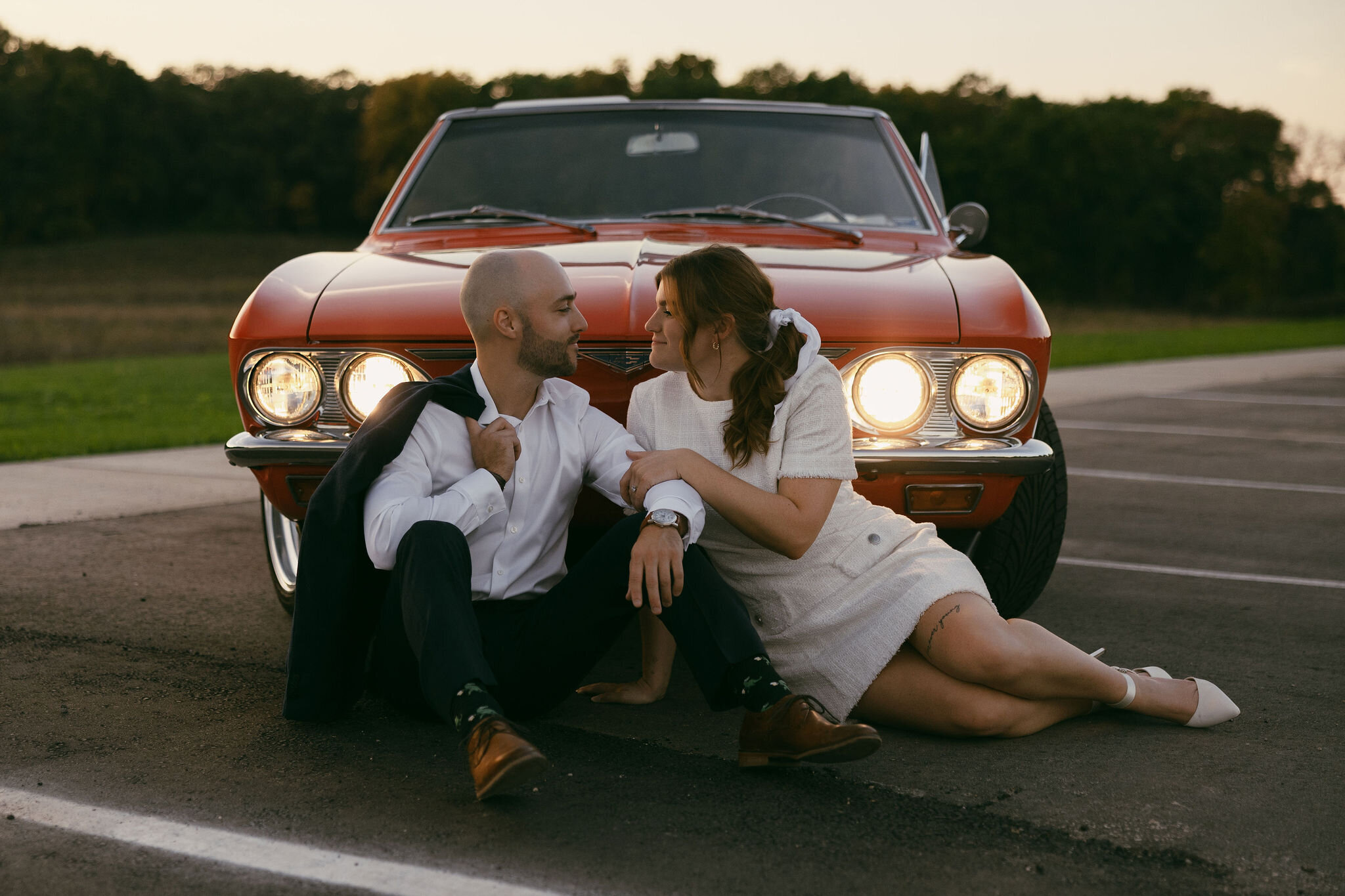 Amazing vintage looking engagement pictures with bride and groom sitting in front of a red classic car with its lights on, looking at each other smiling and touching arms