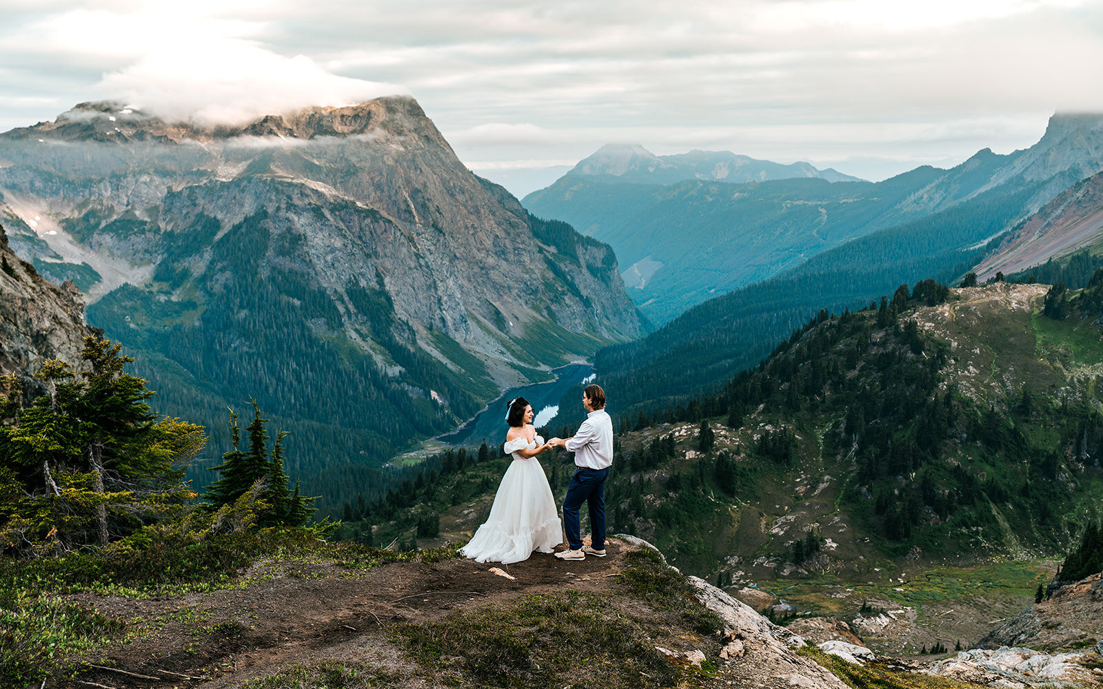 a bride and groom face each other as they exchange vows on the edge of a cliff. blue mountains rise behind them during their adventure elopment