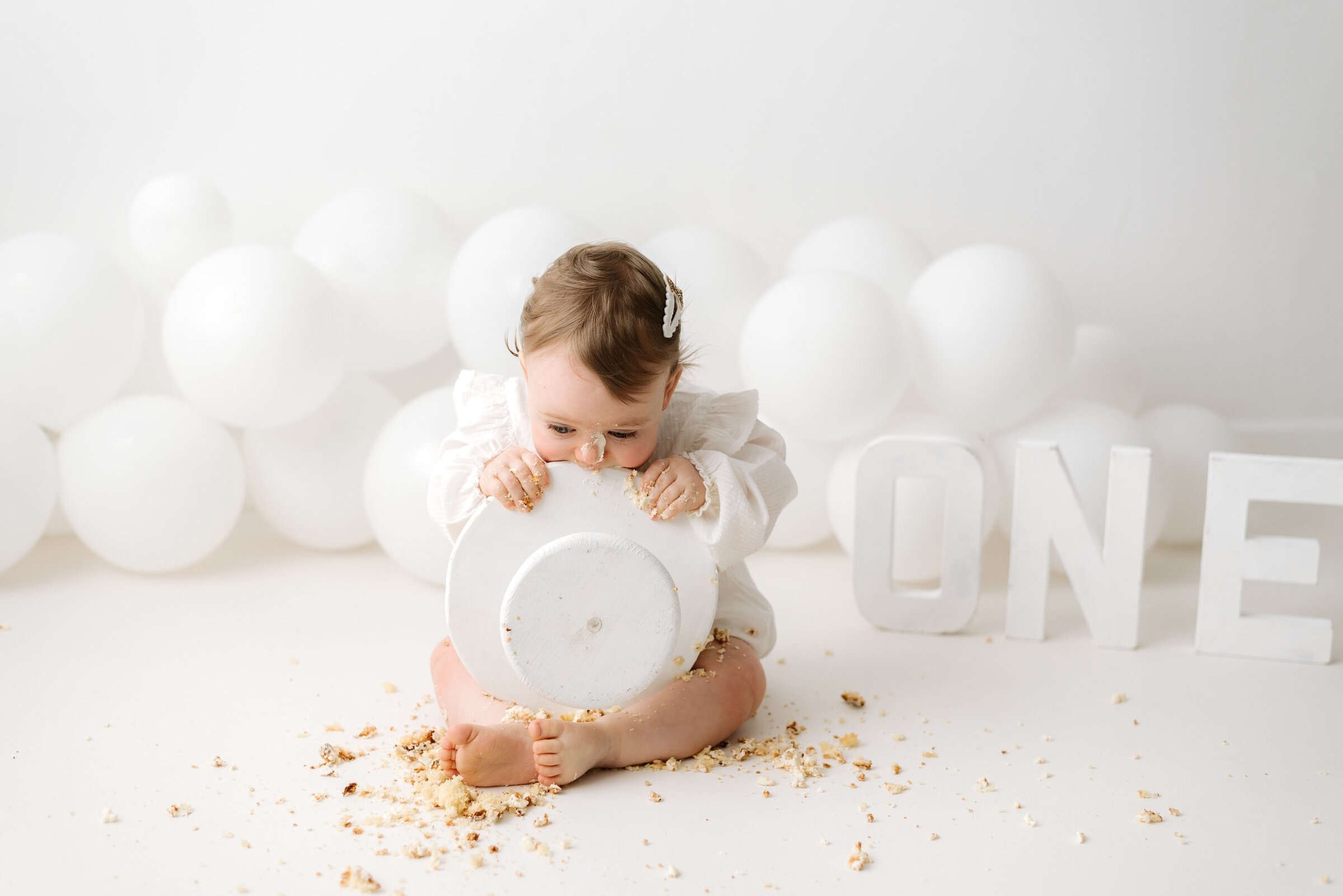 Baby girl sitting in front of balloons chewing on a cake stand at billingshurst cake smash photoshoot