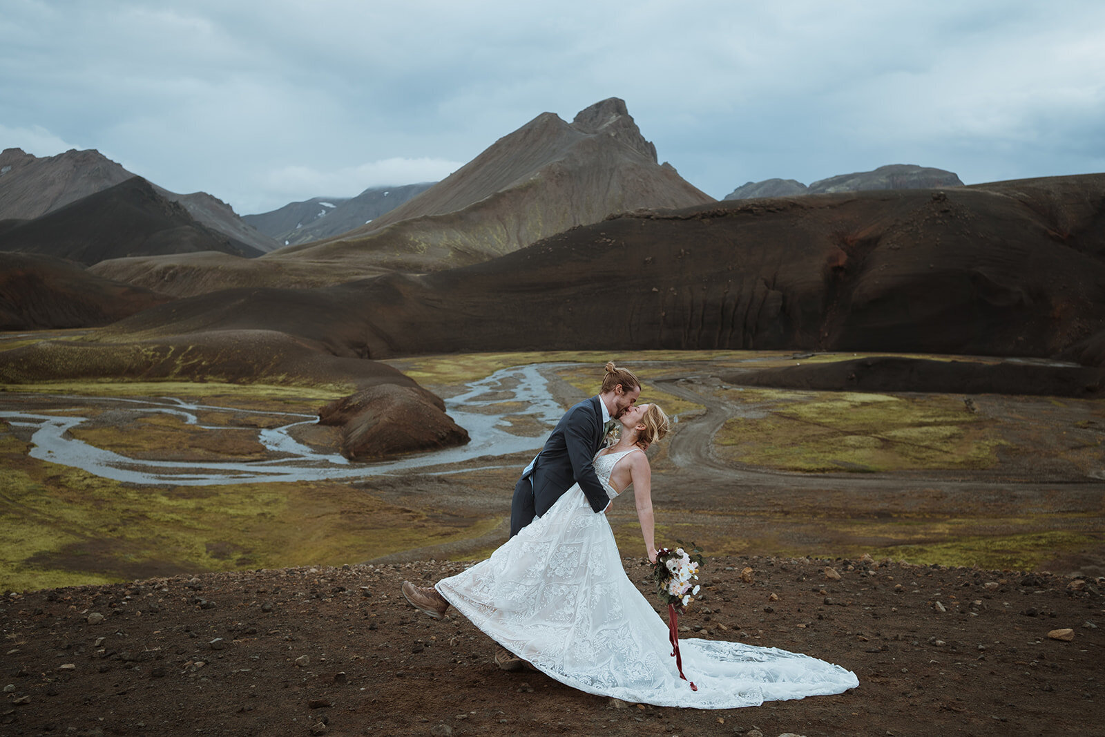the groom is dipping the bride for their first kiss. she has her hand outstretched with flowers. The groom is holding her by the waist and they are overlooking the mountains behind them.