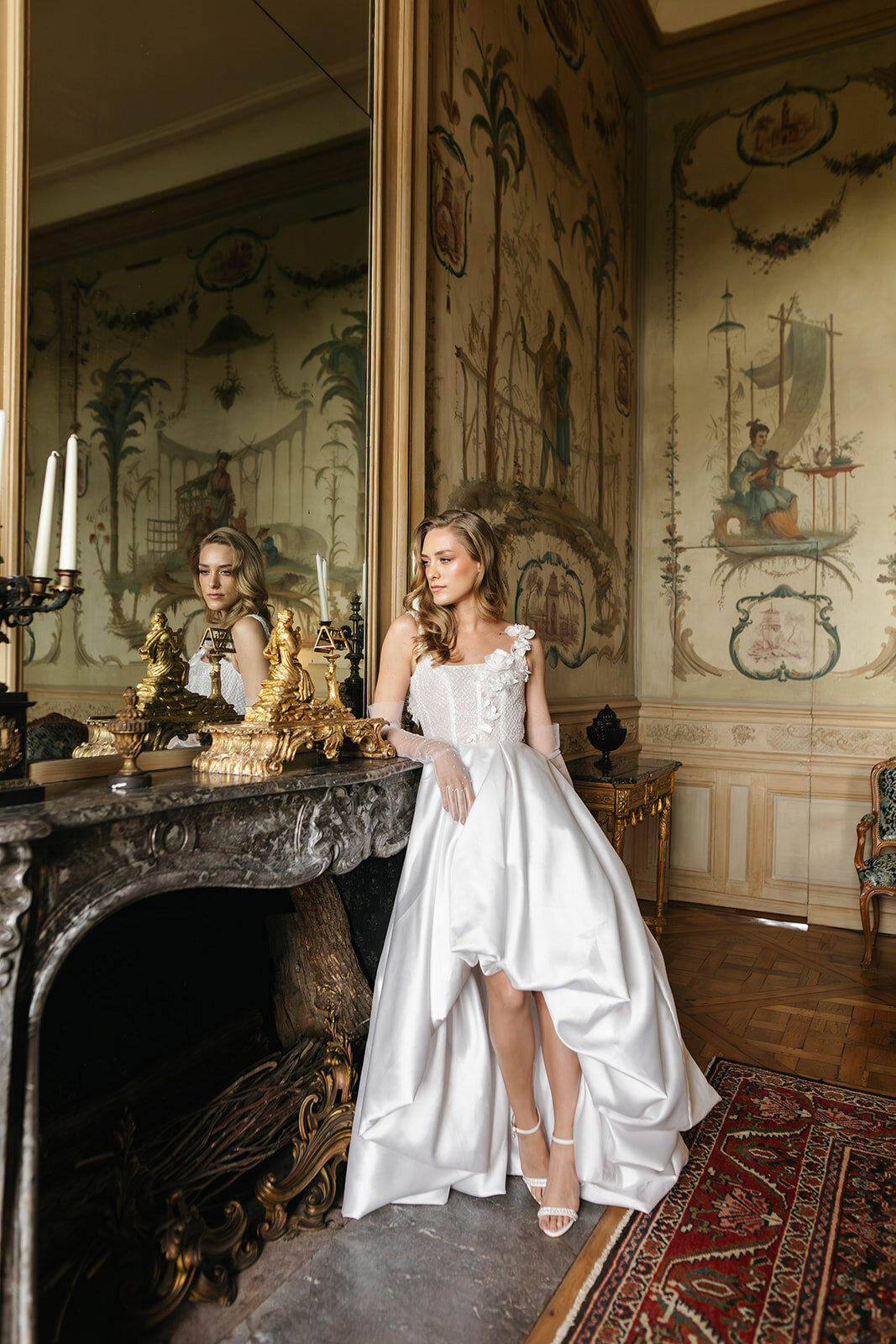 A bride in an elegant white gown stands by an ornate fireplace in a French chateau, surrounded by antique gold decor and painted walls