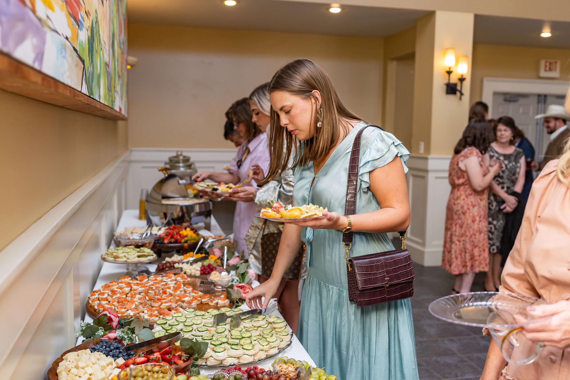 A woman in a light blue dress selects food from a buffet table filled with assorted appetizers, while other people gather in the background at an indoor event.