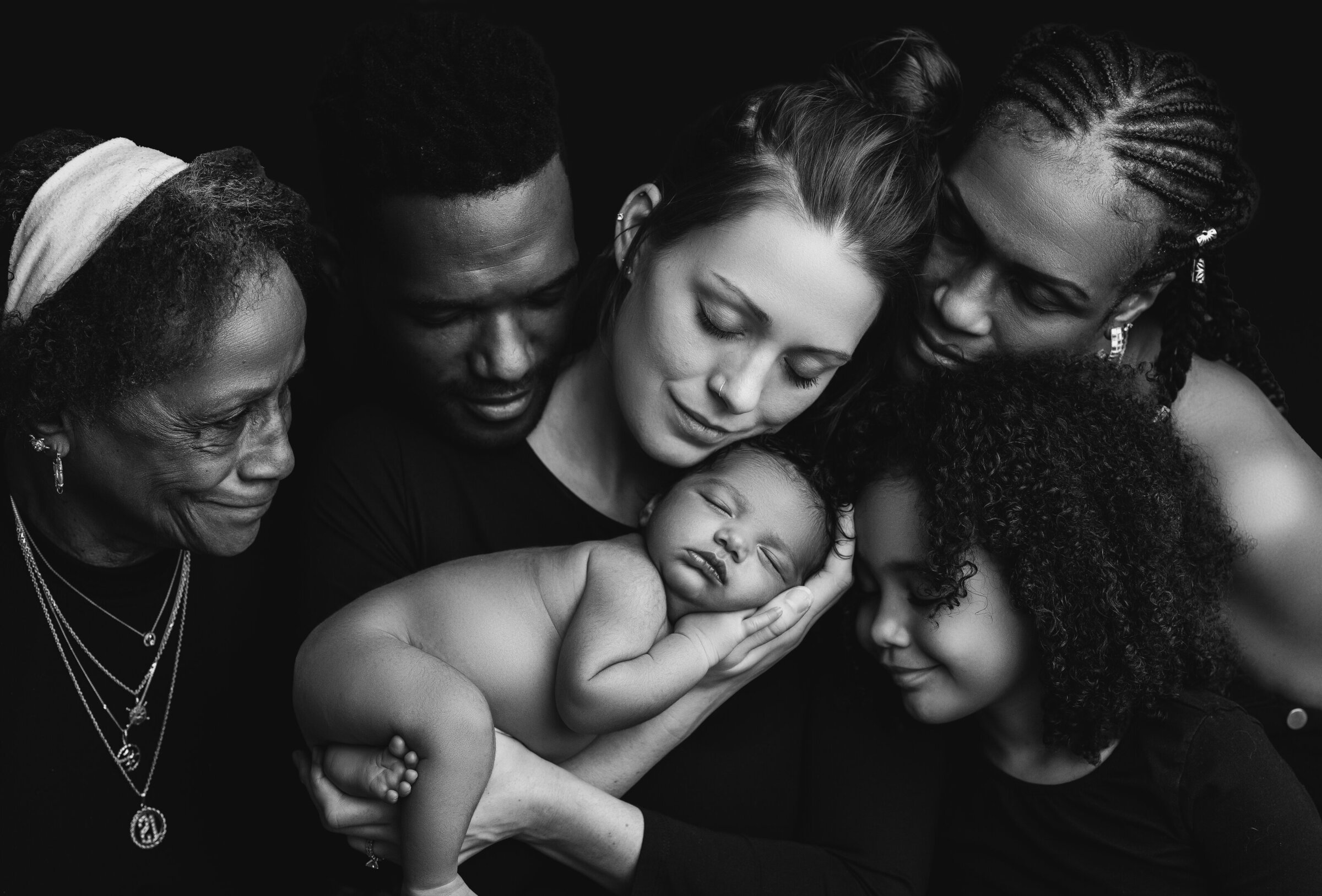 A black and white photo captures a diverse family in Waxhaw, gathered closely with tender smiles. A baby sleeps peacefully at the center, cradled by the group. This Waxhaw newborn photography exudes warmth, love, and a profound connection among all family members.