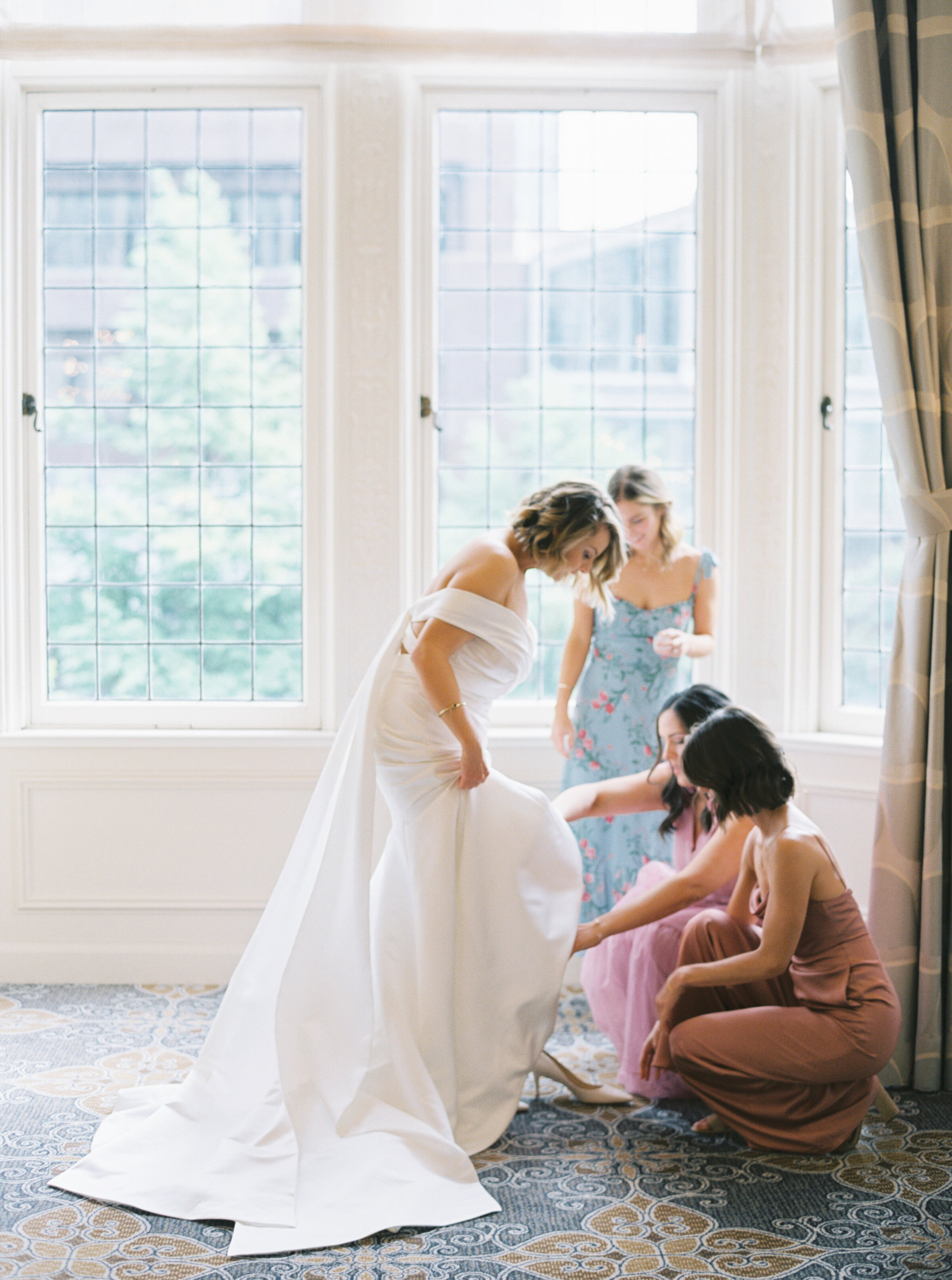 bridesmaids help put the brides shoes on in front of large windows