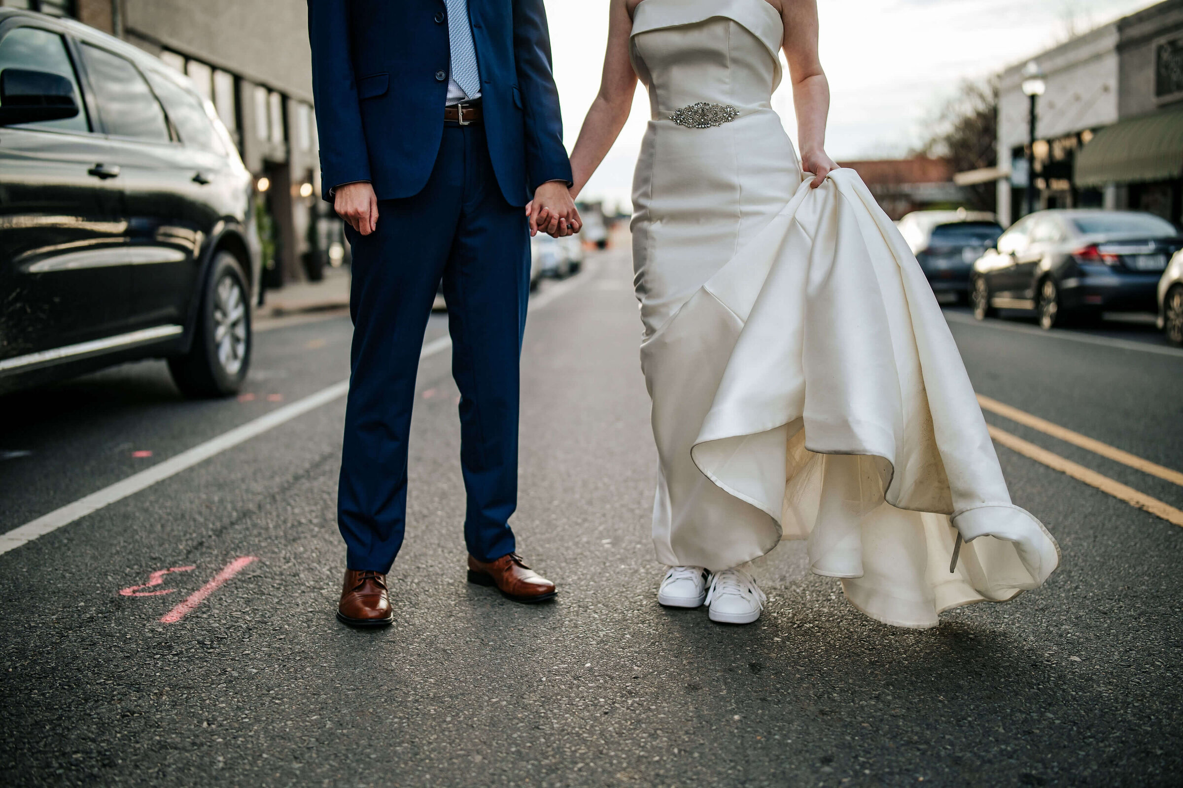 A bride in a white wedding dress and sneakers holds up her gown while standing on a city street, holding hands with a groom in a blue suit. Their faces are not visible. Cars are parked along the street.