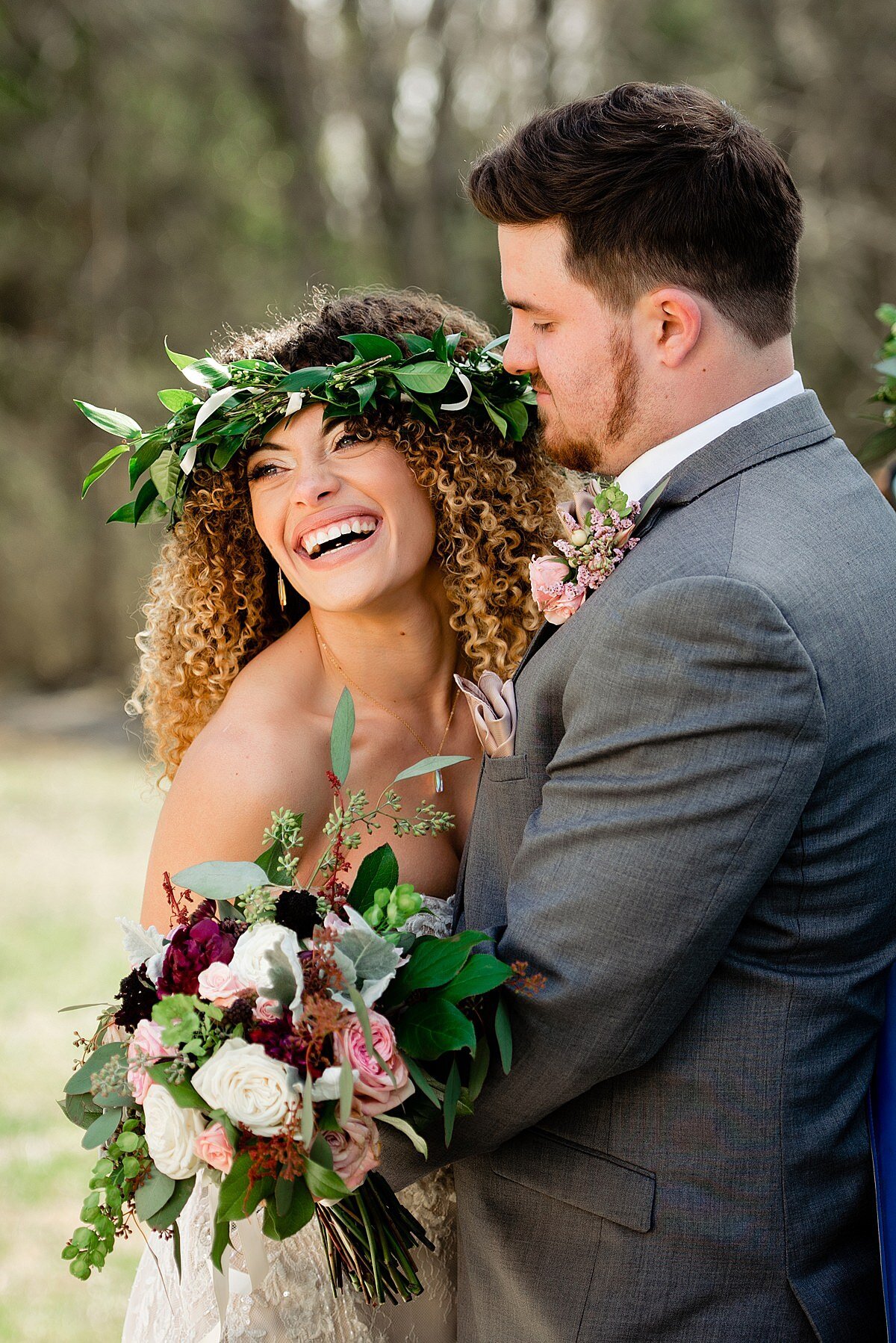 Bride with curly hair wearing  a greenery crown and laughing off into the distance while her husband whispers in her ear