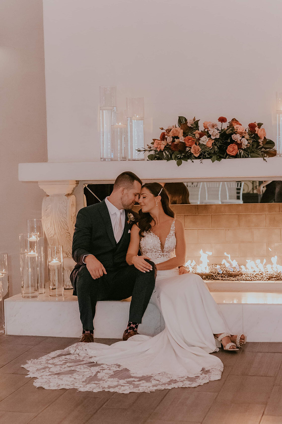 A bride and groom sit closely in front of a glowing fireplace at SoHo63, a Chandler wedding and event venue. Surrounded by candlelight and a floral arrangement in warm tones, the couple shares a quiet, romantic moment in the venue’s cozy and elegant indoor setting.