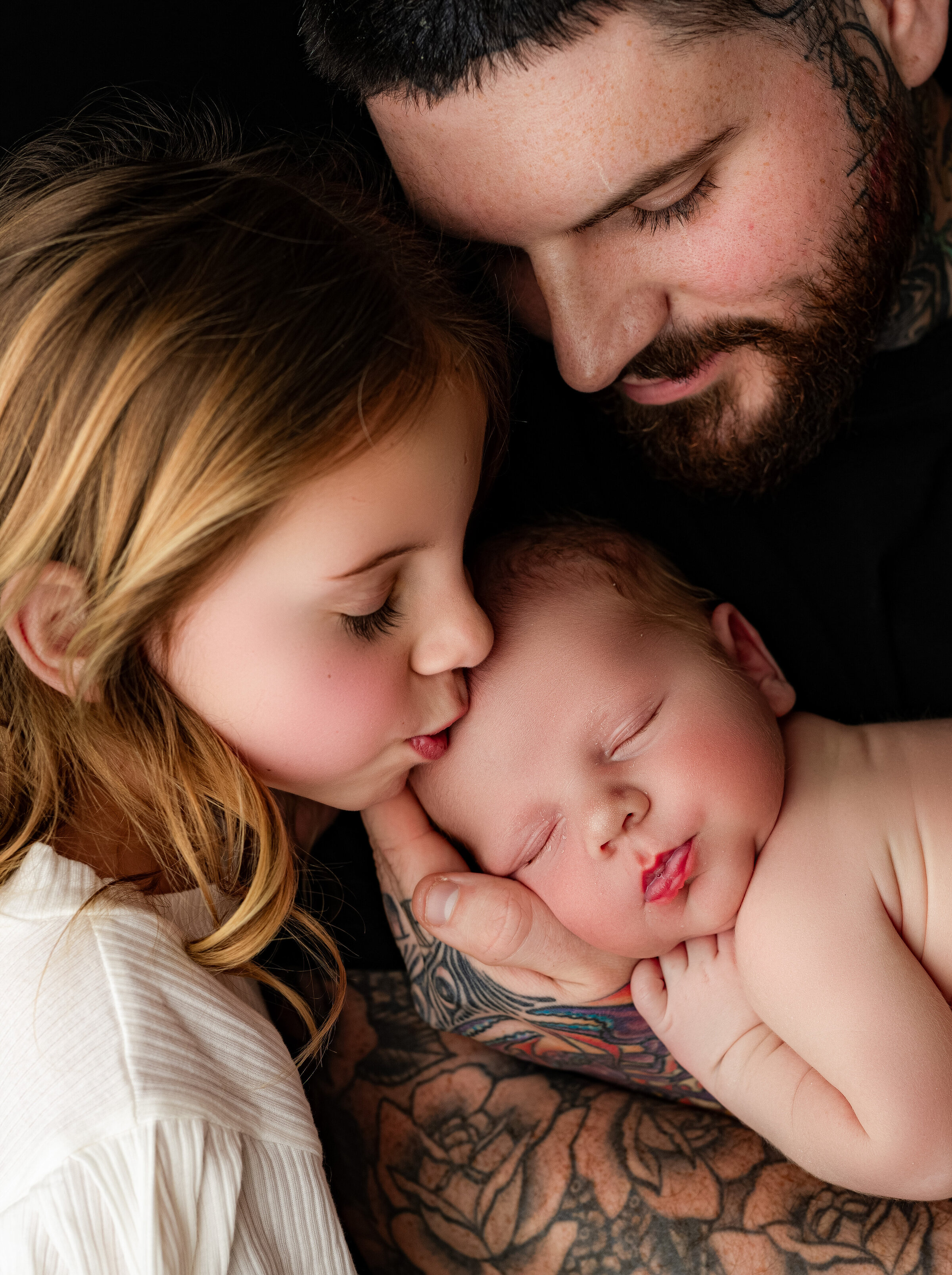 In a tender scene, a young girl gently kisses a sleeping baby on the cheek while a tattooed man cradles the newborn, smiling warmly. Captured by a Waxhaw newborn photographer, this moment overflows with love and warmth.