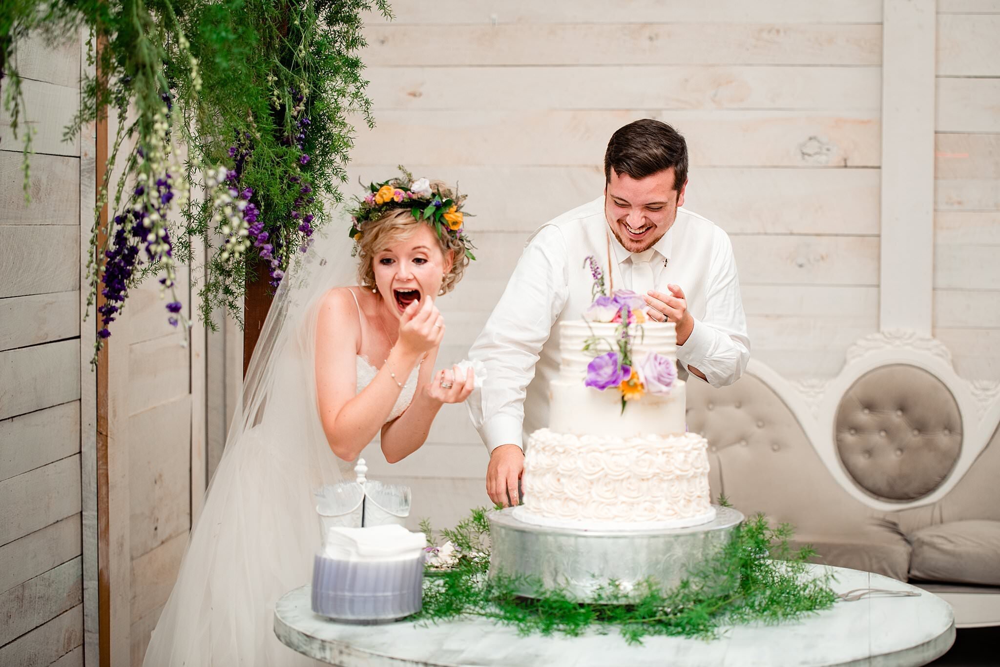 Bride and groom smashing cake into one another faces inside a white barn