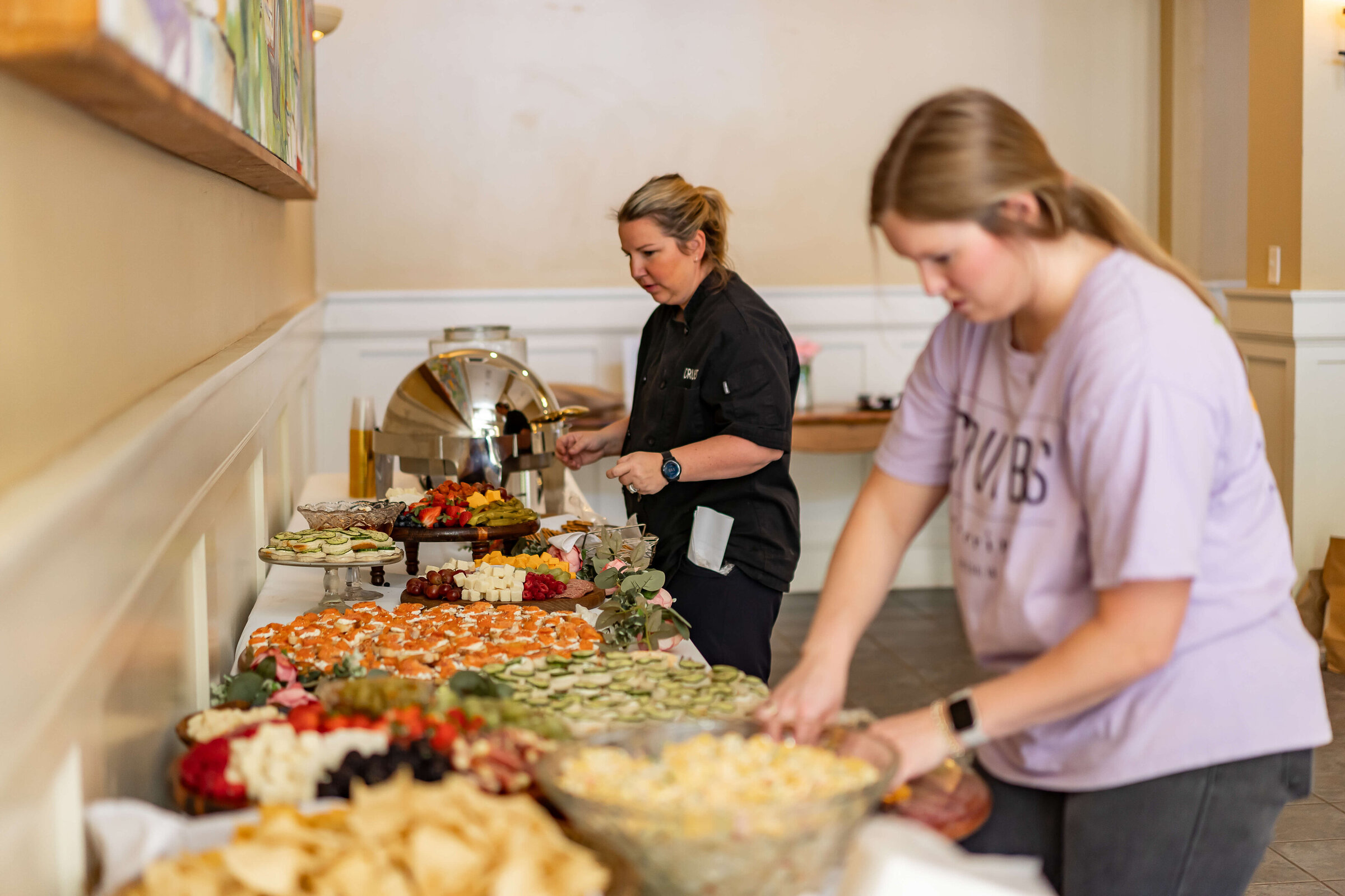 Two women arrange food on a long buffet table covered with various appetizers and snacks, including chips, vegetables, and fruit, in a bright indoor setting.