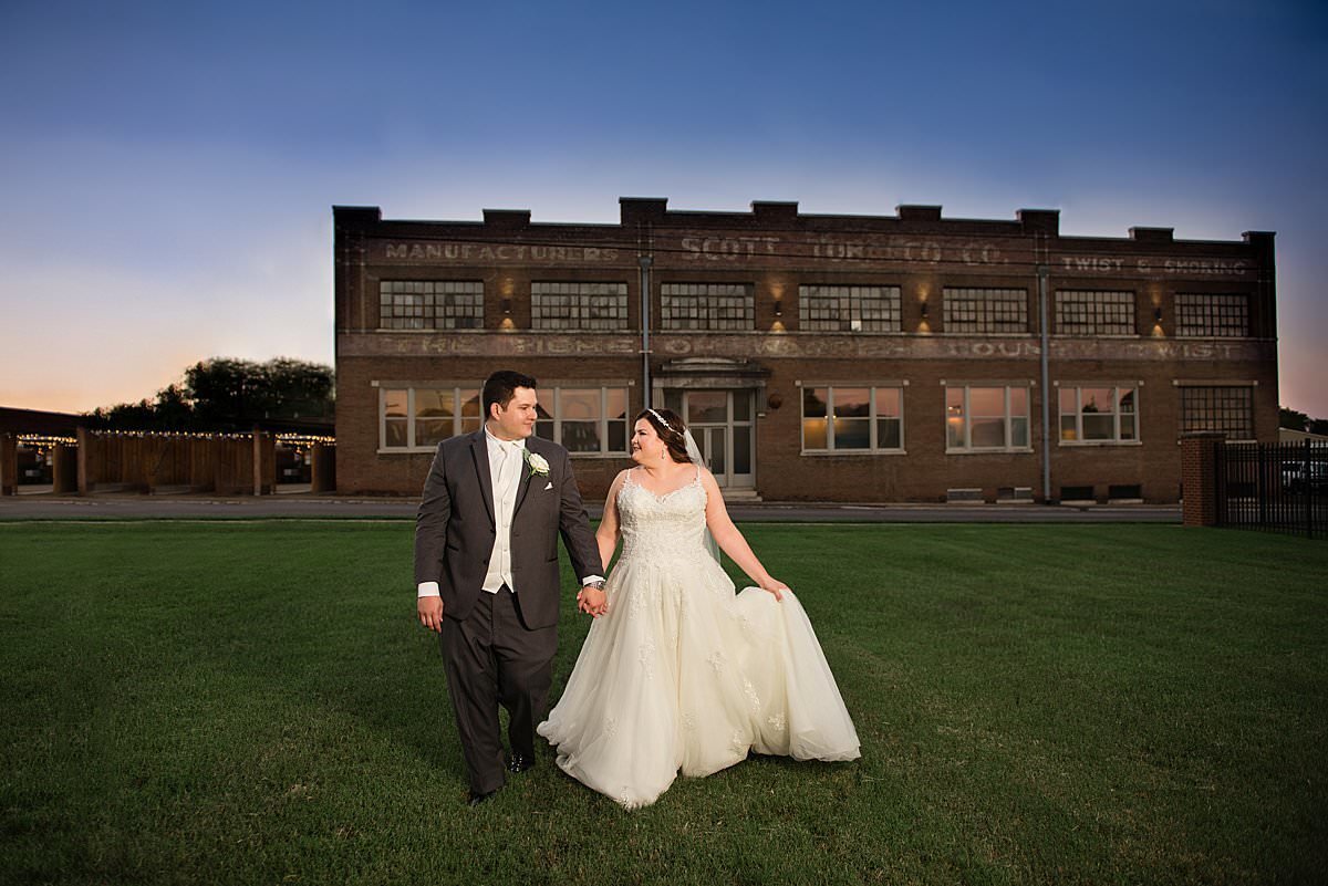 Bride and groom walking through grassy field with the Venue at 939 Adams in the background at sunset