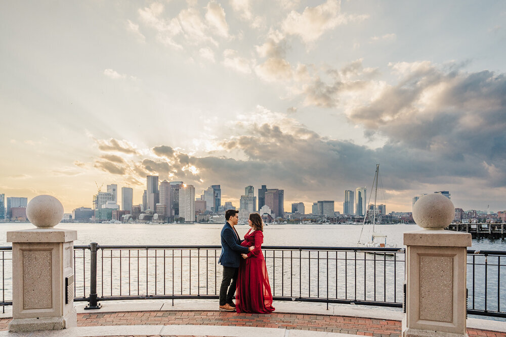 pregnant couple stand face to face at sunset with boston skyline