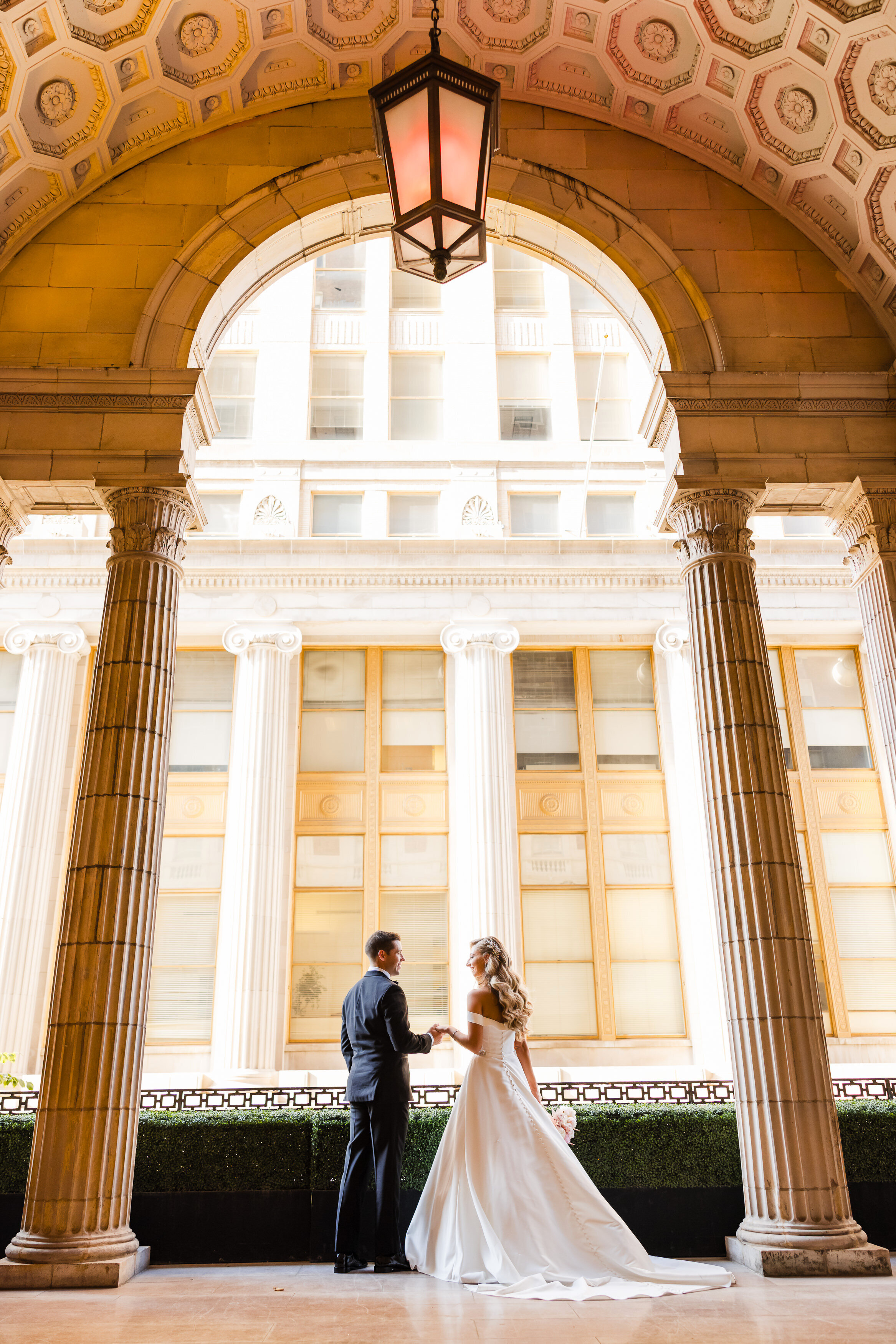 Couple in wedding attire stands gracefully holding hands on the balcony at Ballroom At The Ben