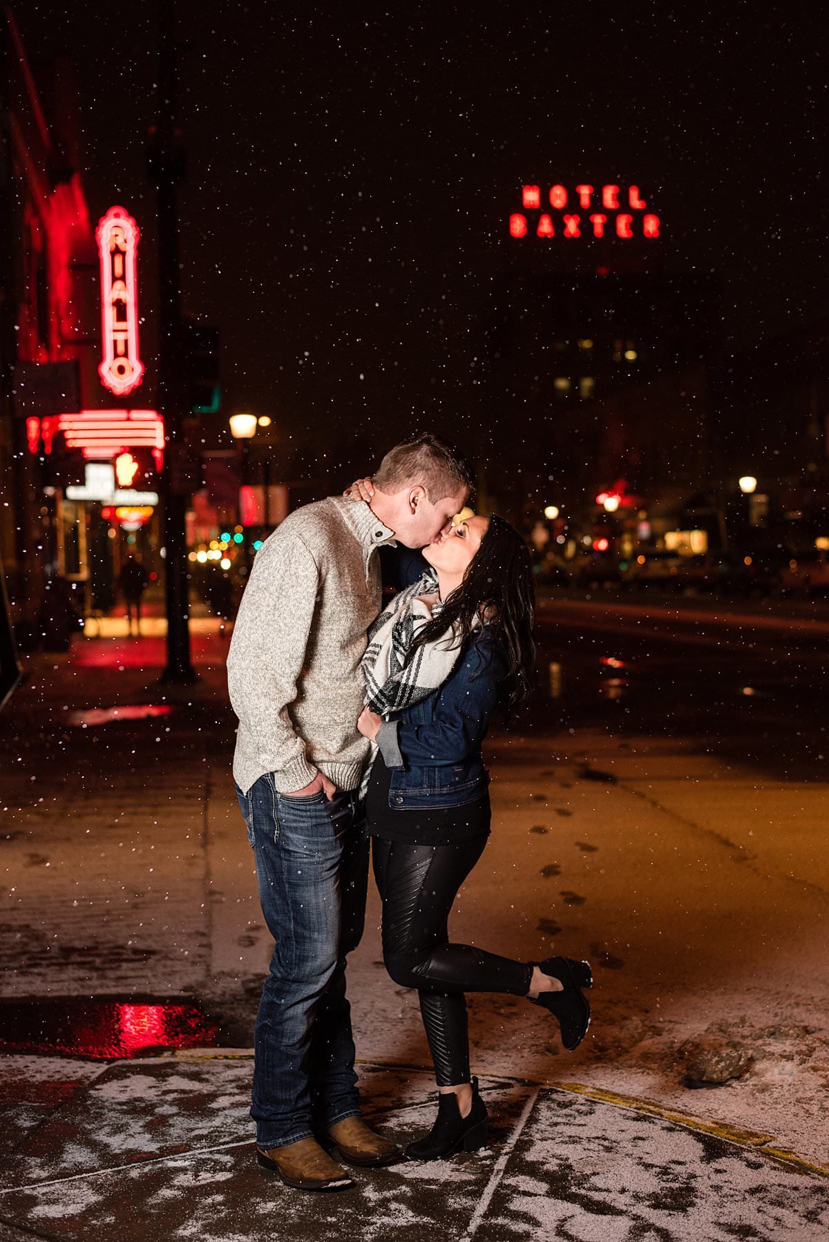 Engaged couple kissing on a snowy night in downtown Bozeman with the Hotel Baxter and Rialto behind them