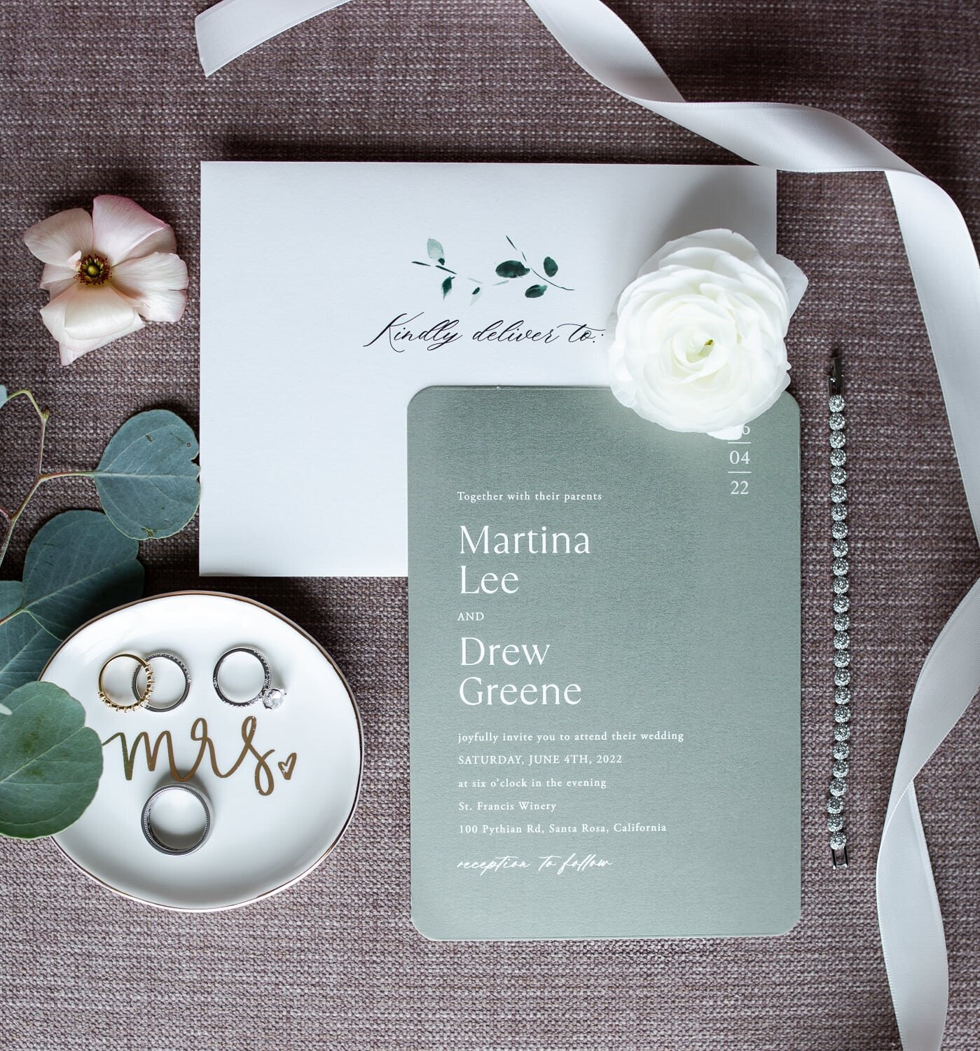 Flatlay, sage and white wedding invitation, white ribbon and bridal jewelry.