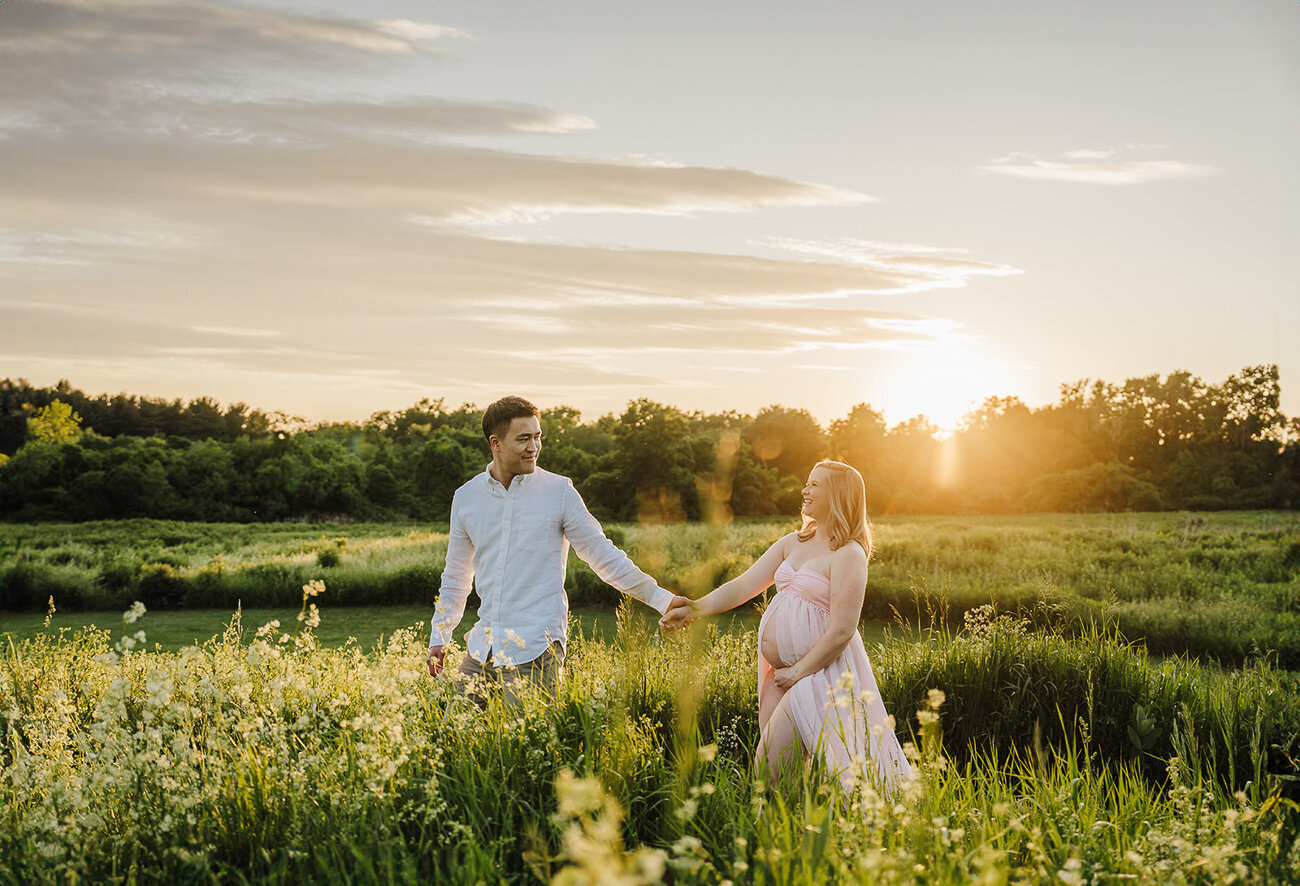 pregnant couple walks hand in hand in sunset field during boston maternity photos