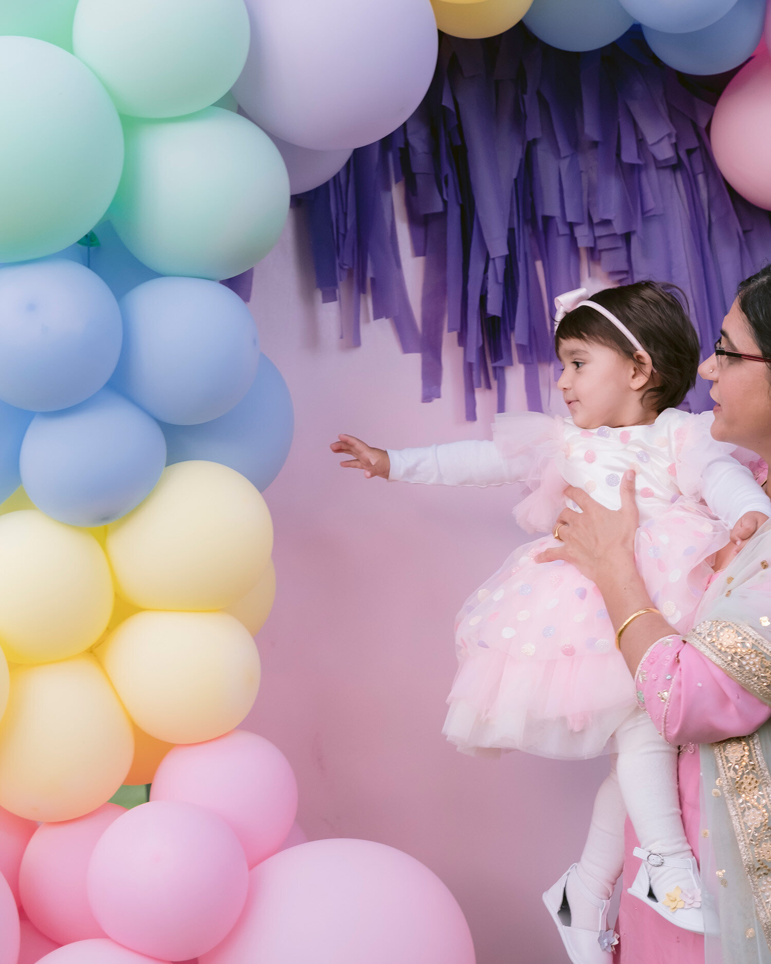Bay Area birthday photographer captures baby reaching for a balloon arch while being held by her nanny during a first birthday party