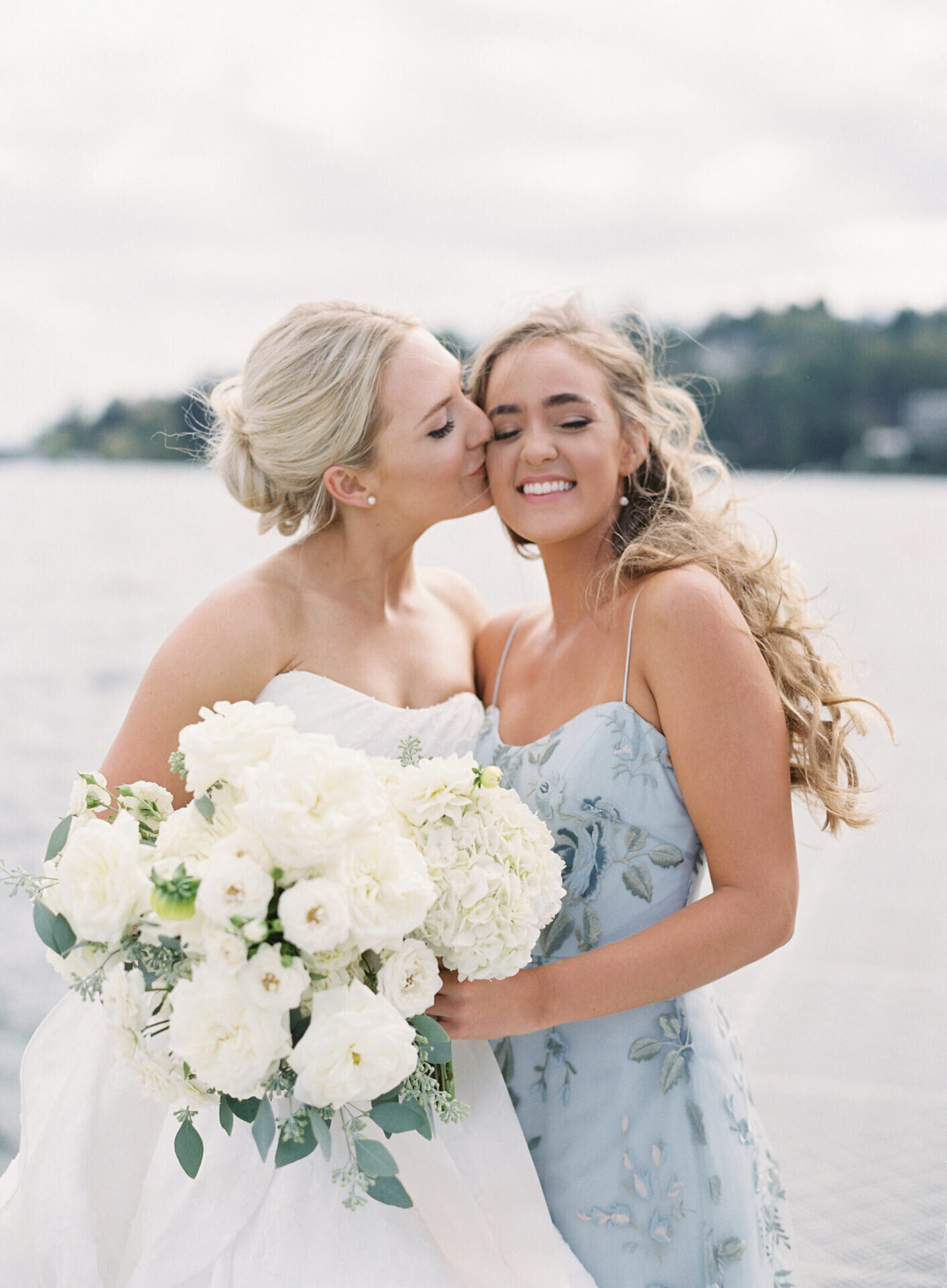 bride holding lush white rose boquet while kissing the cheek of her smiling bridesmaid