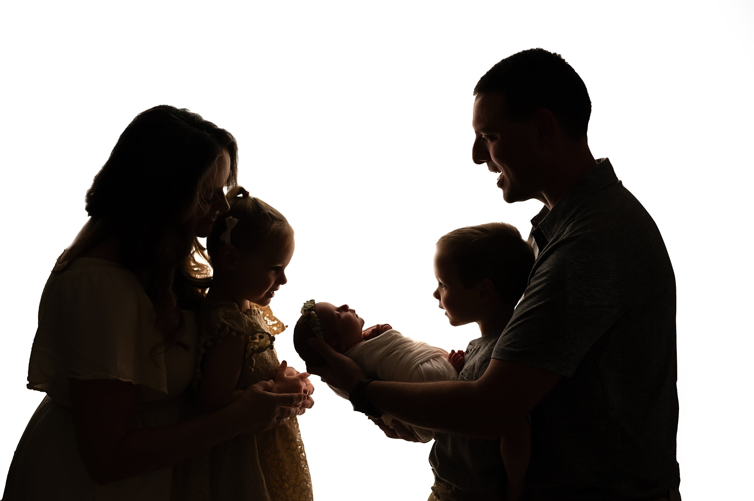 Silhouette of a family of five against a white background captures the warmth and affection as a man cradles a baby, while two young children and a woman lean in. Perfect for those seeking cherished Charlotte newborn photography moments.