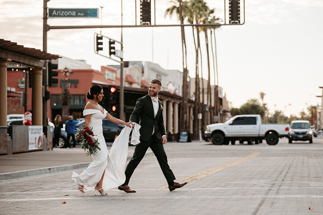 A bride and groom cross Arizona Avenue in downtown Chandler, just steps from SoHo63, a Chandler wedding and event venue. The bride holds a bold red bouquet as the groom helps lift her dress, capturing a fun, fashionable wedding moment in the heart of Chandler’s historic district.