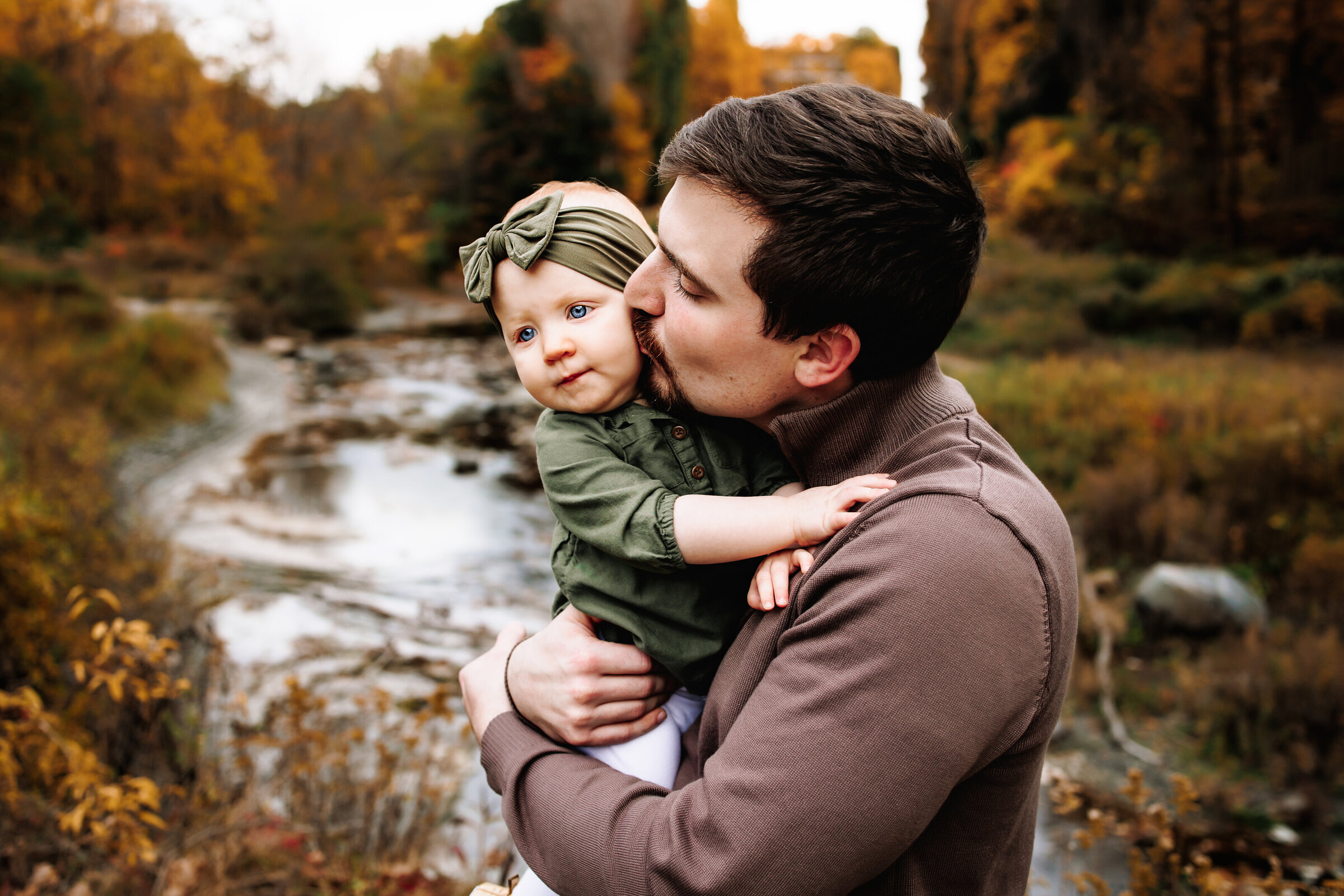 Father gives his baby girl a kiss on the cheek amongst a backdrop of a stream lined with fall color