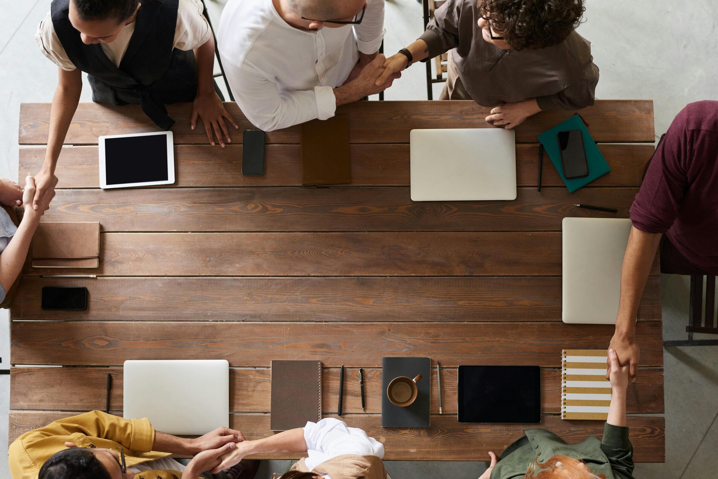 Image of an aerial view of a conference desk with people surrounding the desk having a meeting, with notebooks and coffee on the table