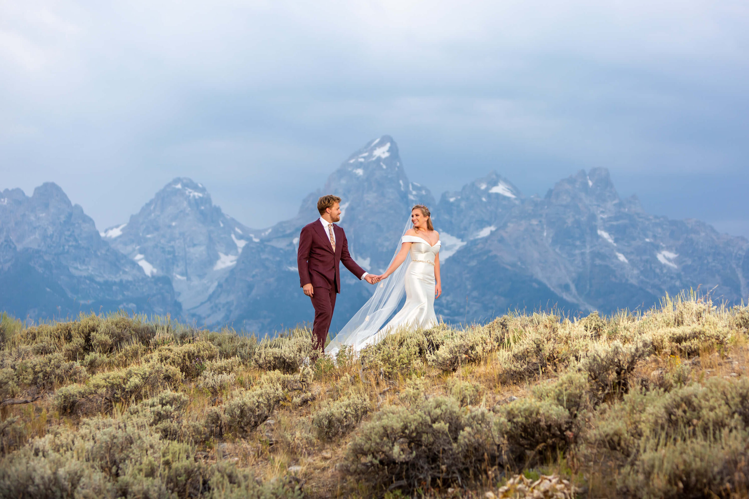A newlywed couple walks hand in hand across a sagebrush-covered hillside in Grand Teton National Park. The bride’s flowing veil catches the wind, and the groom looks at her with admiration. Towering mountain peaks frame the backdrop for this dramatic wedding moment.