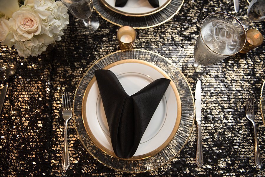 Black, silver and gold sequin tablecloth overhead photo of table setting of glass charger and white and gold plate with black  napkin