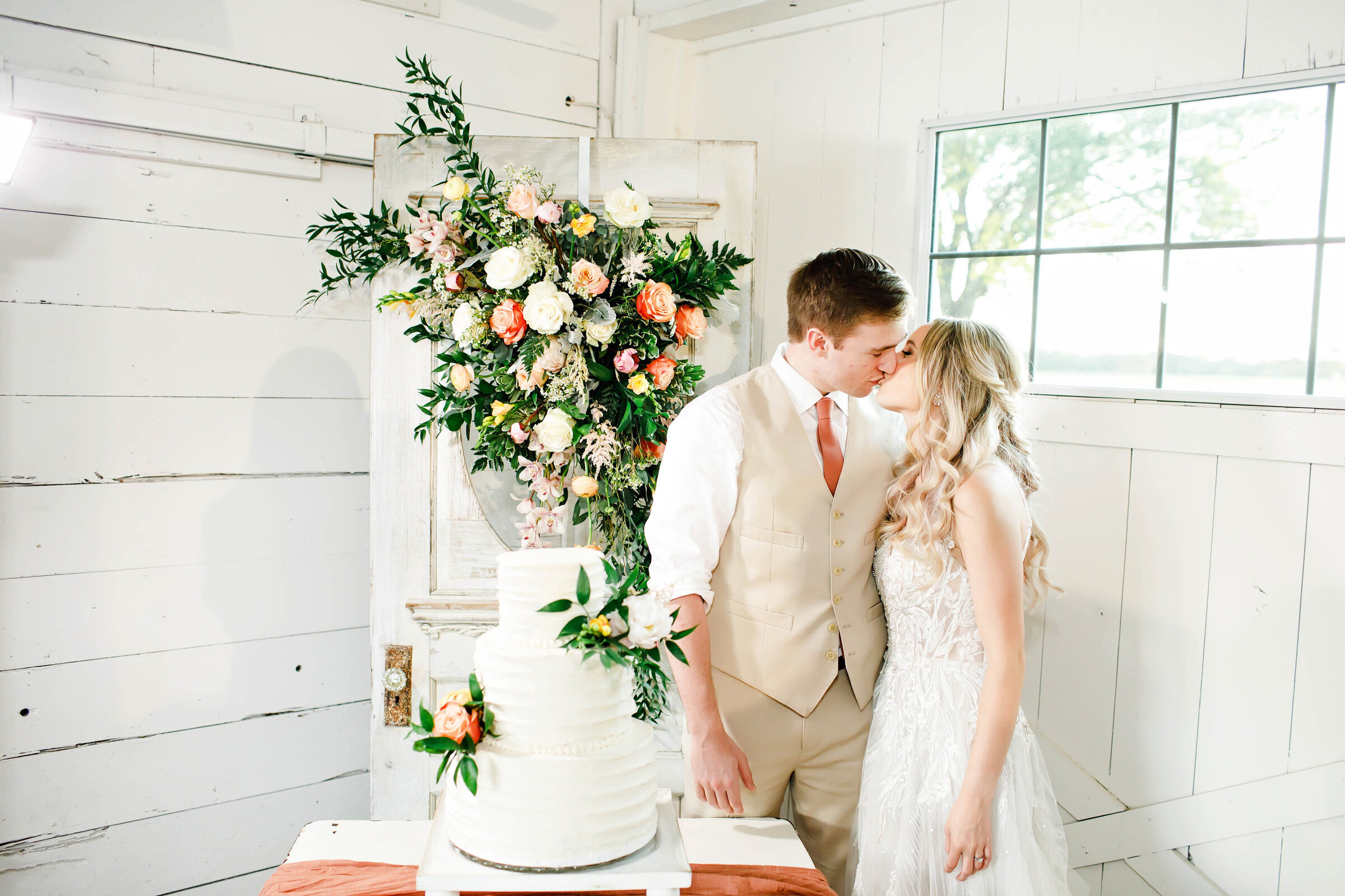 A bride and groom kissing each other by their wedding cake. Behind them is a gorgeous floral arrangement and an all white wedding cake with florals on it