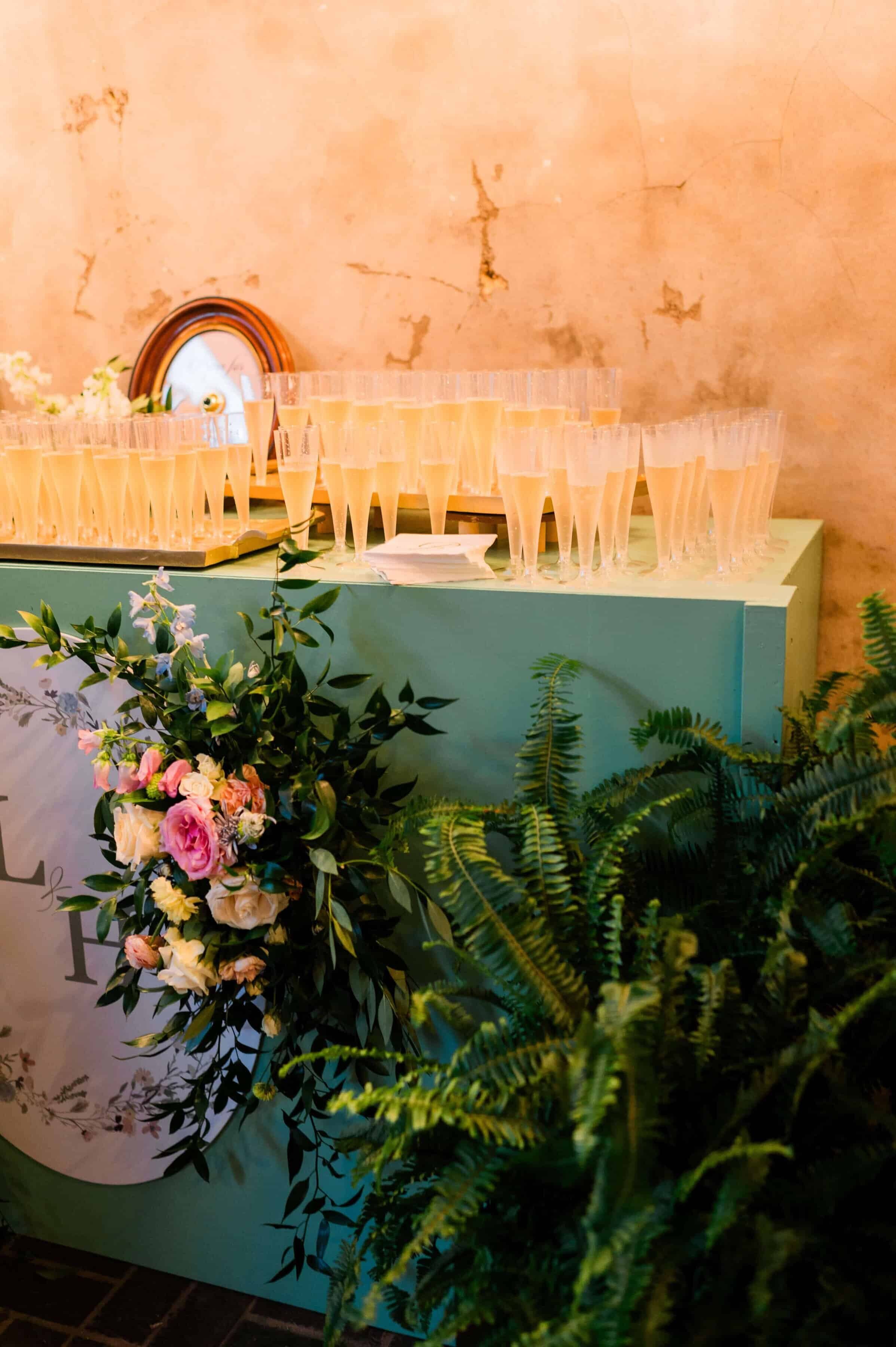 A green table decorated with a floral arrangement and ferns holds numerous filled champagne flutes at a private event. Behind the glasses is a round clock against a textured, beige wall.