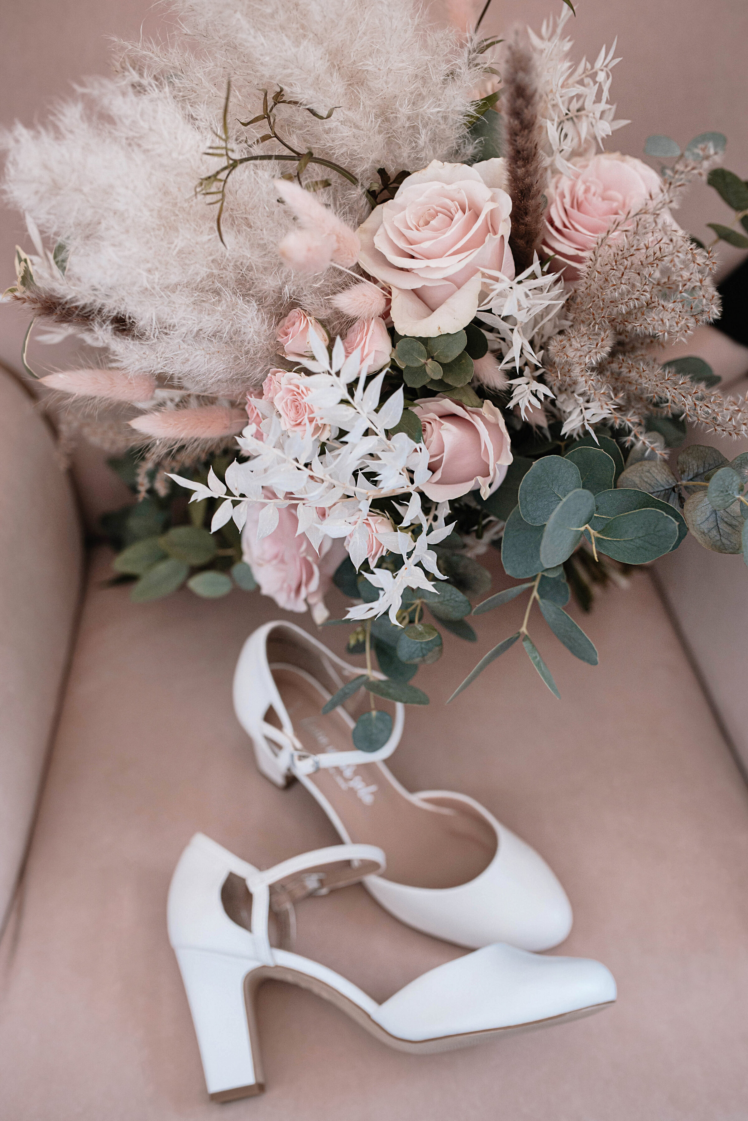 Bridal shoes seated on velvet pink chair with bridal bouquet of roses, bunny tails and eucalyptus
