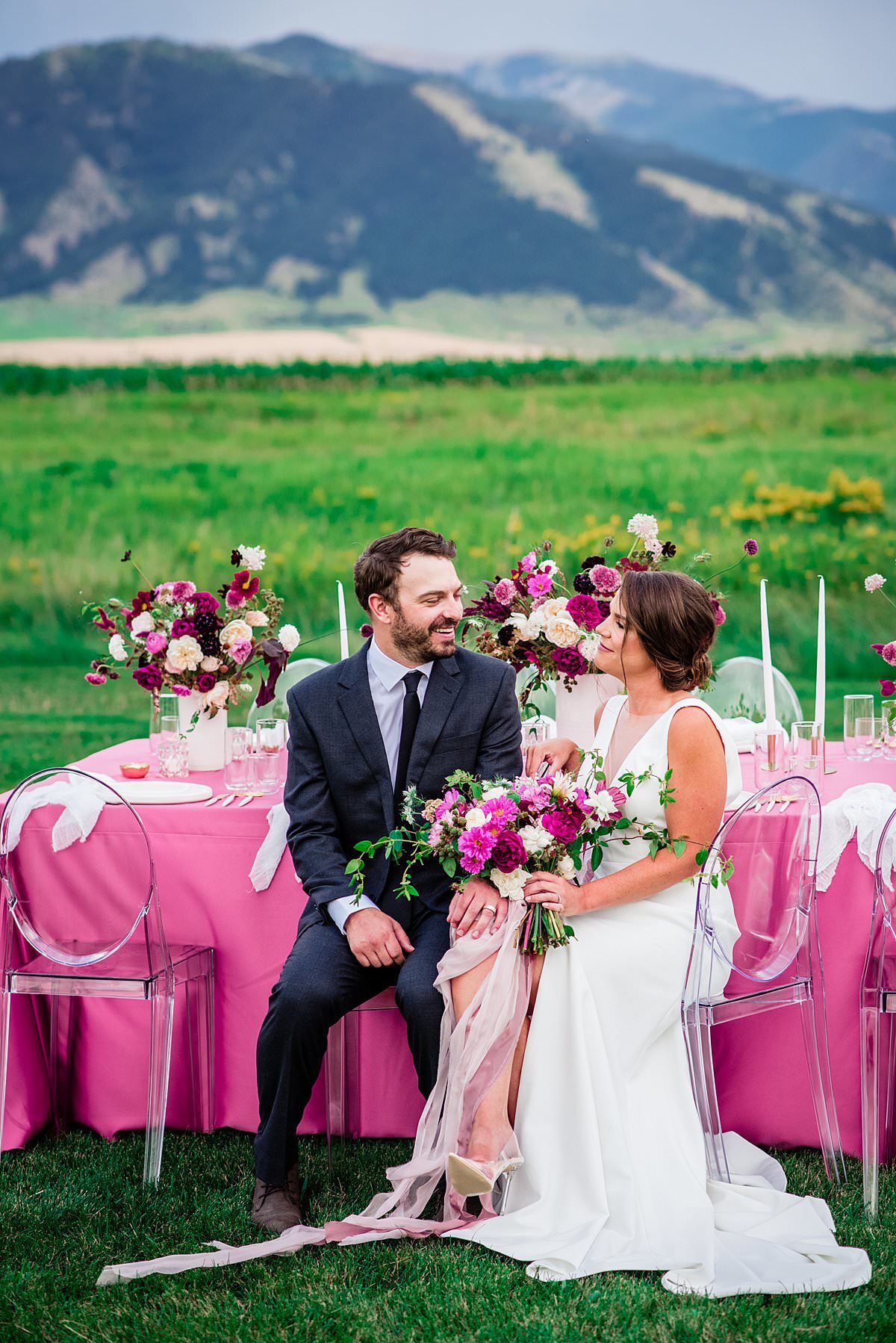 Hot pink and burgundy wedding designs with contemporary acrylic chairs outdoors in a field with Montana mountains  behind thee couple who are sitting together at the table