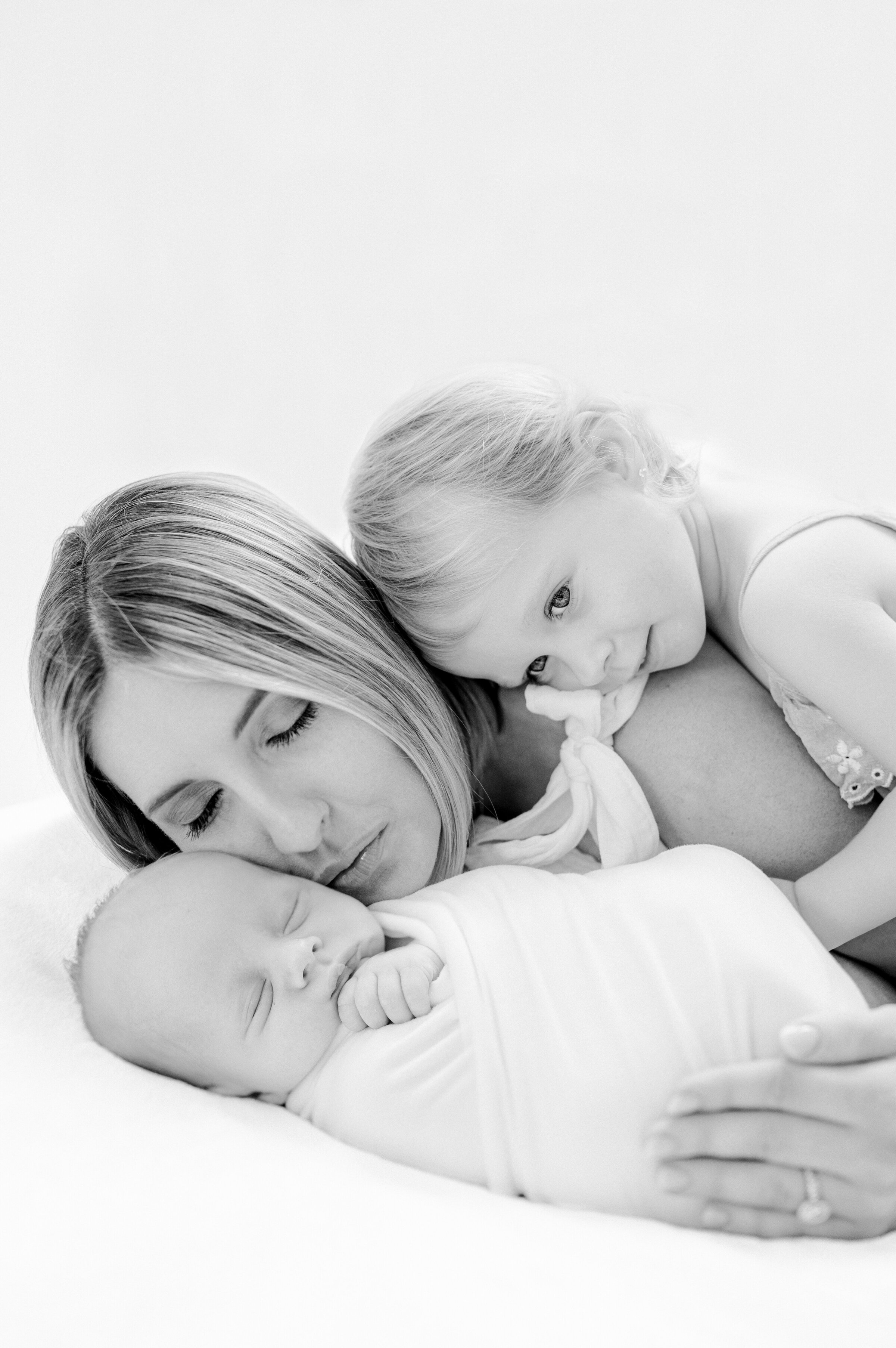 Black and white portrait of mom hugging her newborn and the toddler is hugging mom