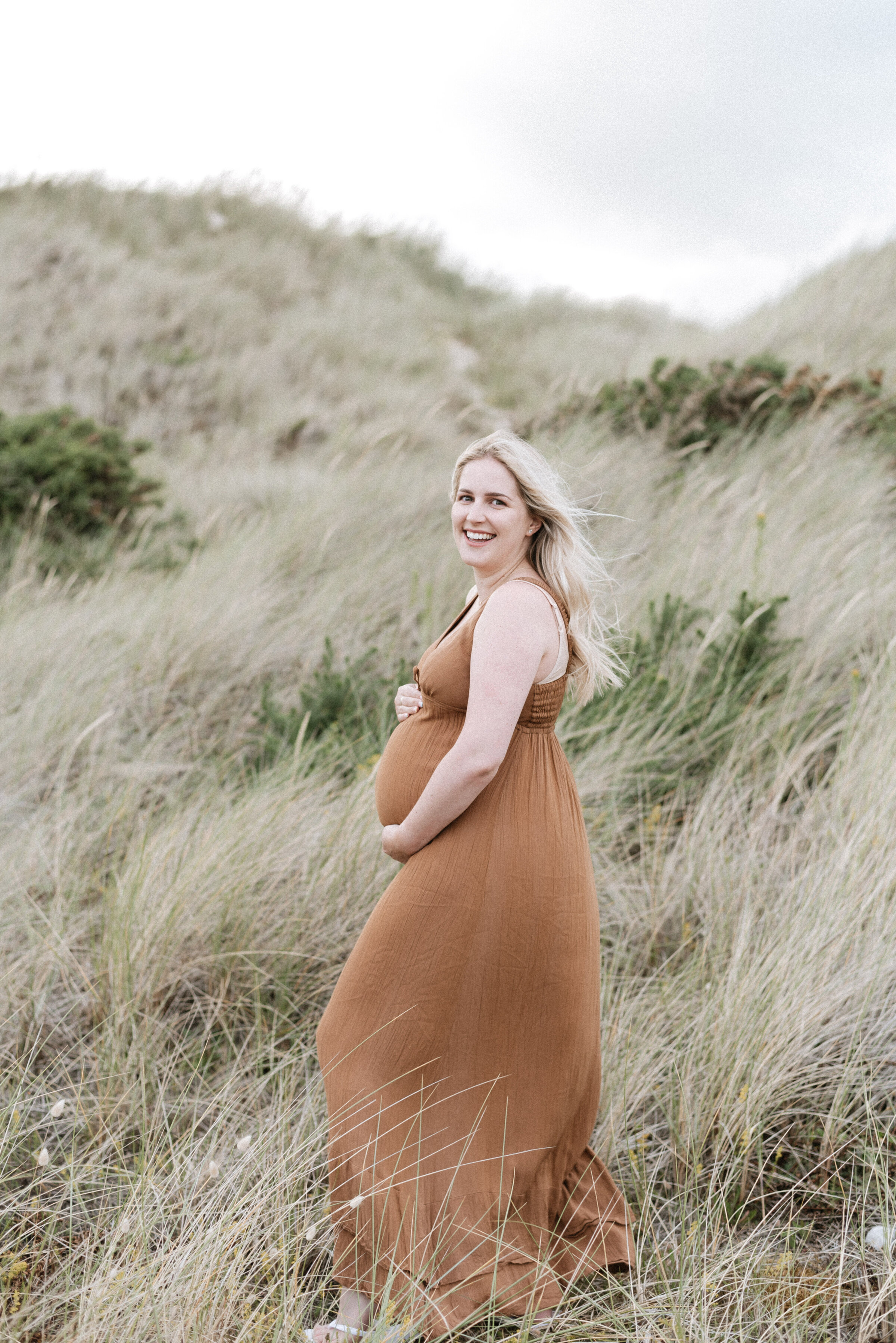 Mother holding pregnant belly smiling  standing on dunes at the beach at West Sussex maternity photoshoot