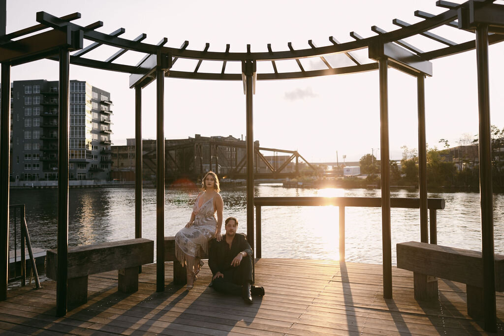 Sunset engagement photos taken on the river in Milwaukee, WI with bride sitting on a wooden bench, groom sitting in front of her on the deck, her hand on his shoulder