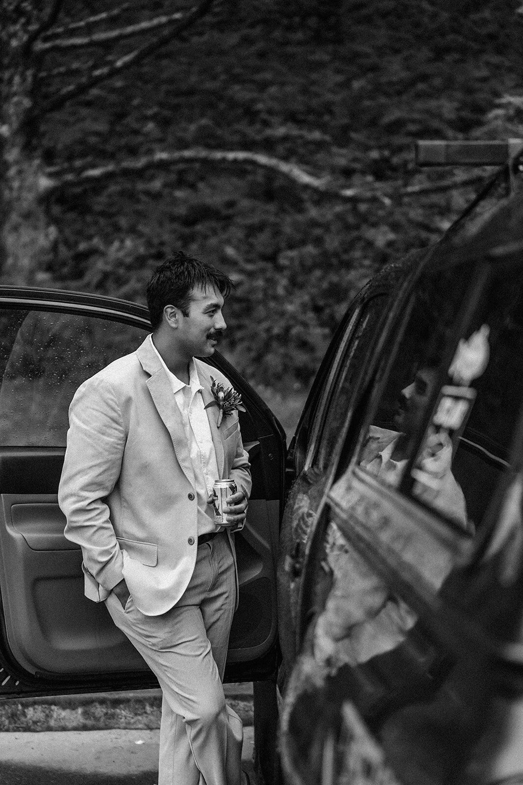 a black and white photo of a groom in his wedding attire. He leans against an open car door talking to his bride as a sips a beer, resting his other hand in his pocket