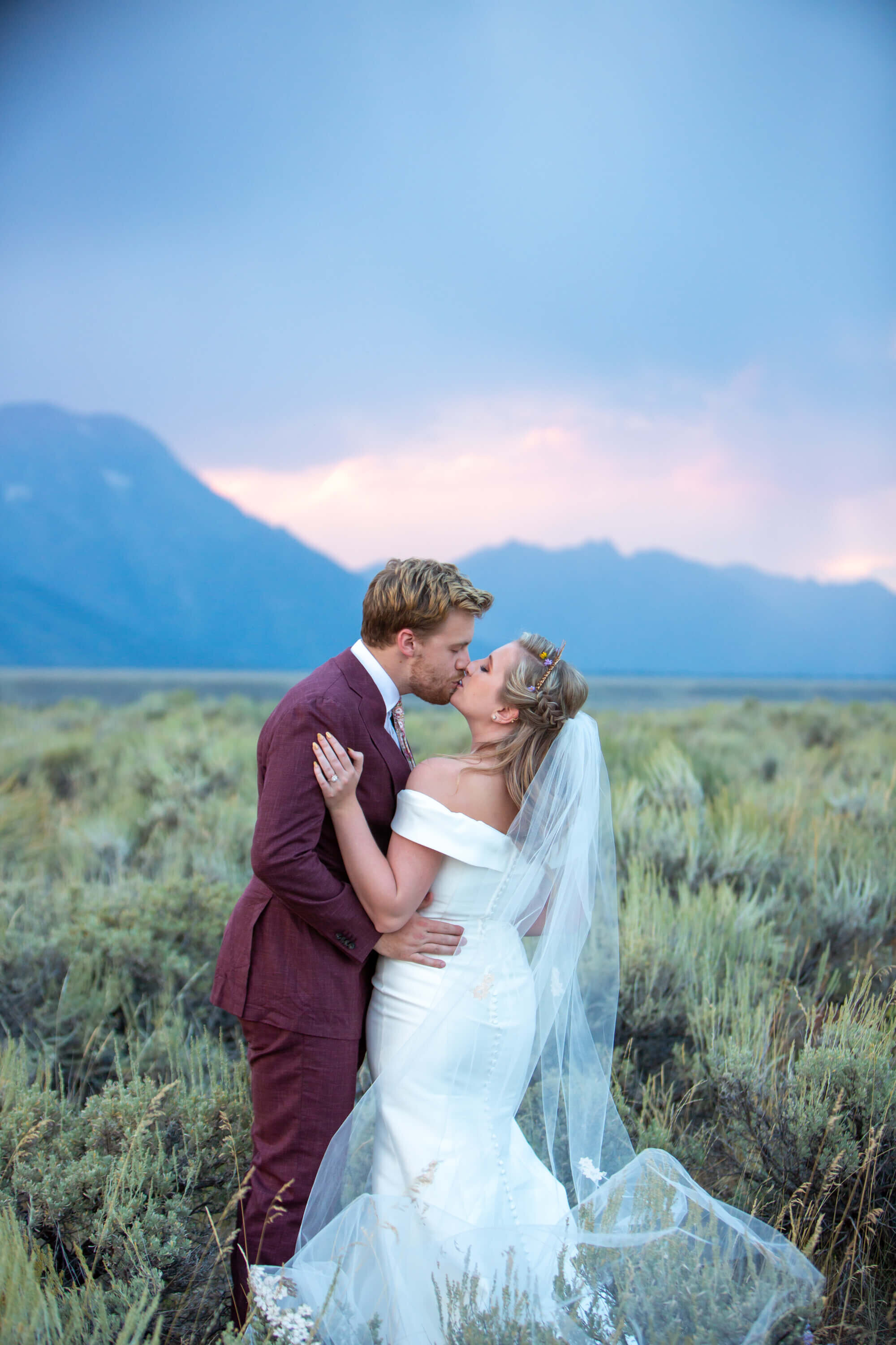 A newlywed couple in a sagebrush field in Grand Teton National Park, with soft pink and blue hues in the evening sky. The bride wears a white off-the-shoulder wedding gown with a flowing veil, and the groom wears a burgundy suit. Their wedding day moment is framed by the Tetons in the background.