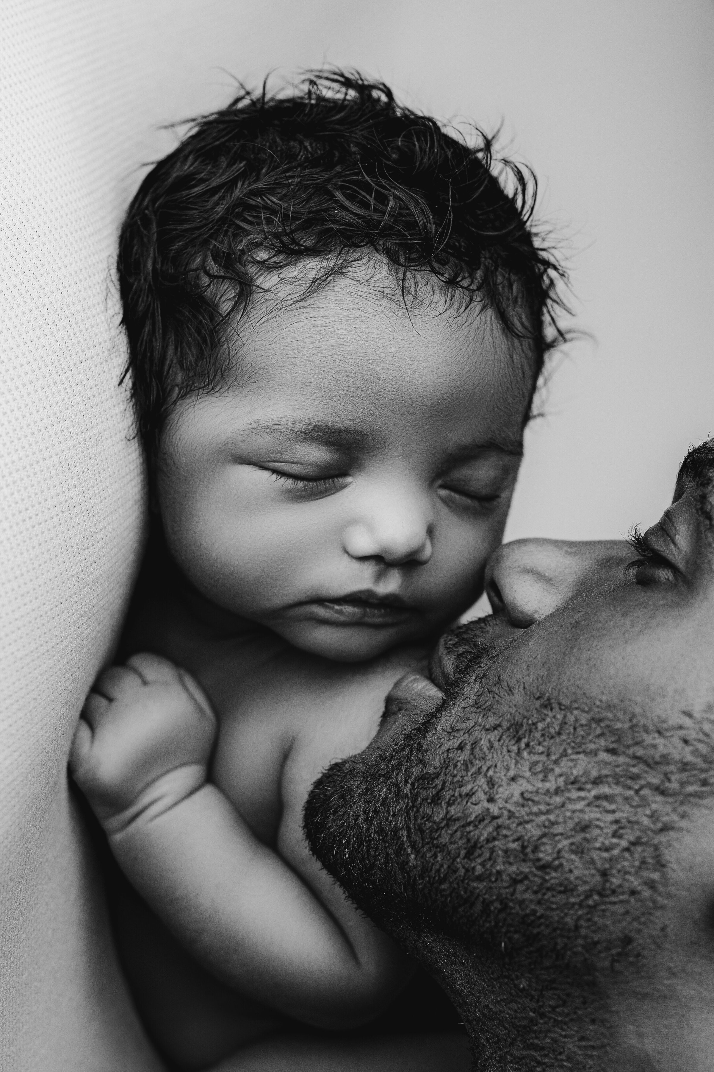 Black and white image of a father tenderly kissing his asleep newborns cheek, captured in the style of Charlotte newborn photography. The baby rests peacefully against a cushion, eyes closed, while the fathers loving expression conveys deep affection and warmth.