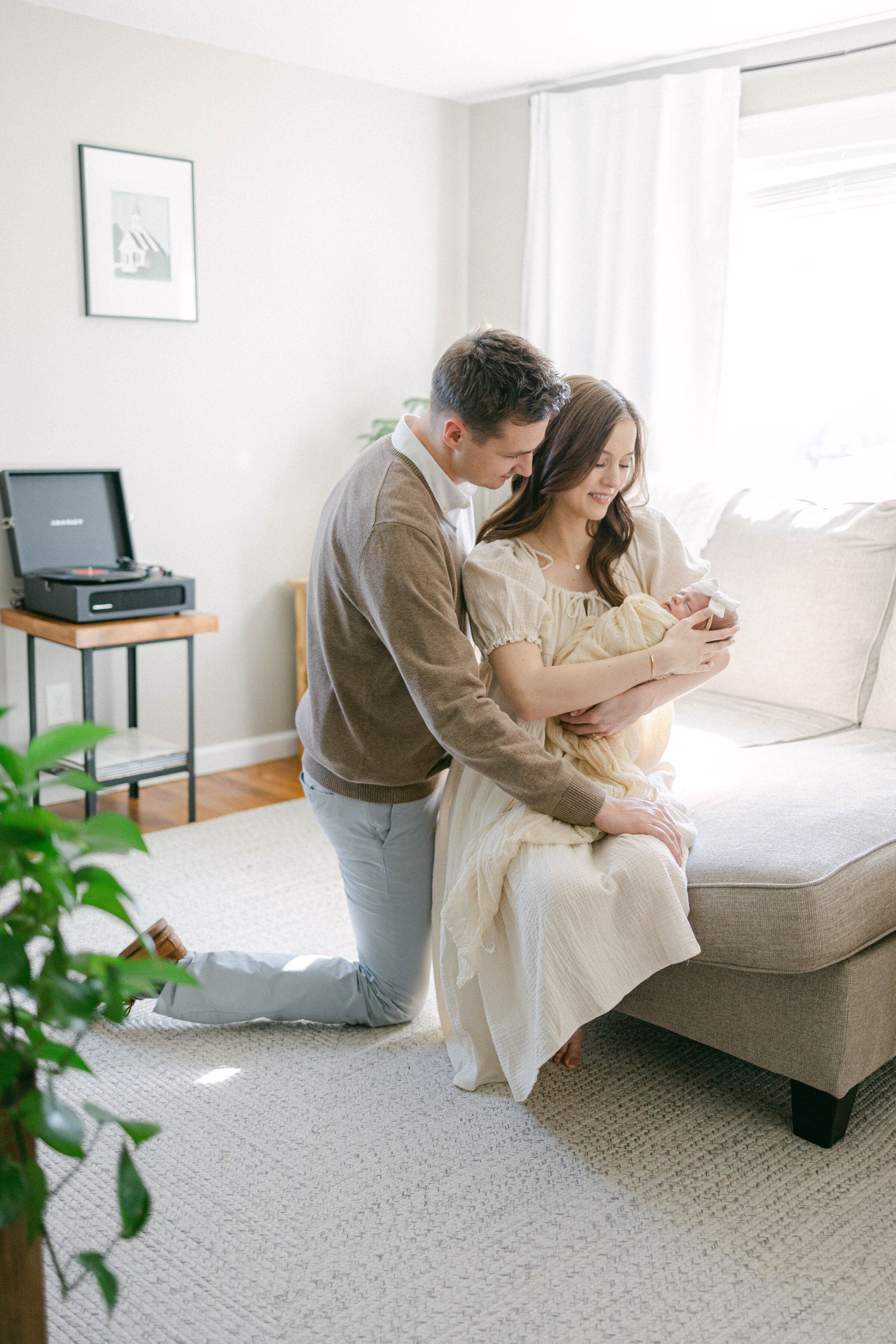 women sitting on couch holding newborn baby with man kneeling on the floor with arms around her and looking at baby knoxville tennessee newborn photographer