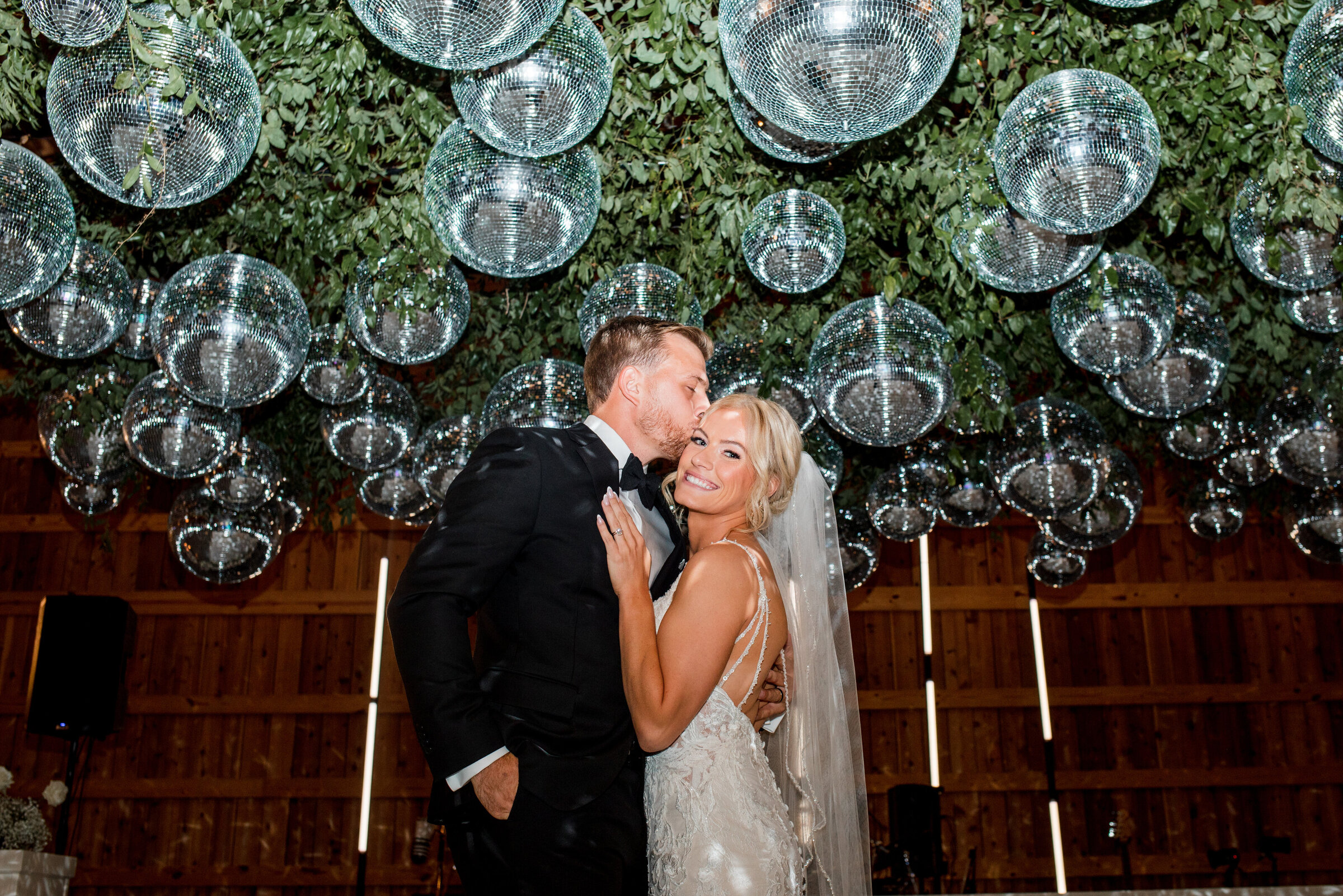 Groom kissing bride during their reception under their greenery and disco ball covered ceiling